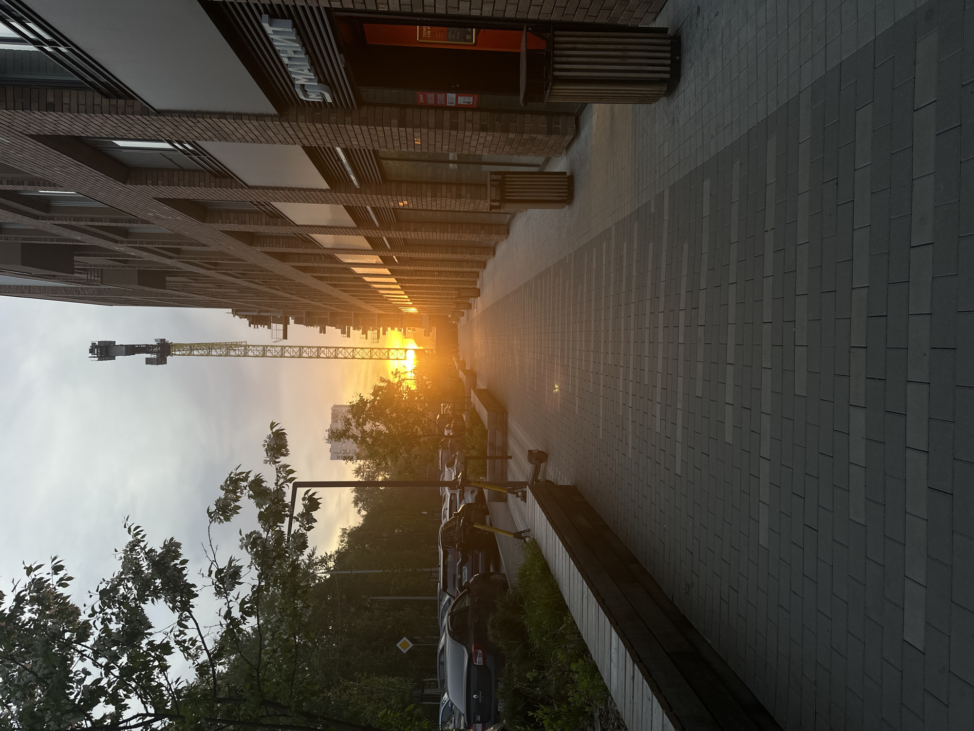 The image shows a street scene at sunset in Yekaterinburg, Russia. A modern brick building with the sign "МАГНИТ" is on the left. A walkway paved with rectangular gray stones stretches into the distance, converging at the horizon where the sun is setting. A construction crane is visible against the sky near the center of the image. On the right side of the walkway, there are wooden benches and parked cars. Trees and other greenery line the street on the right. The sky is a mix of light gray and orange due to the sunset. There are people present, although they are small and difficult to distinguish.