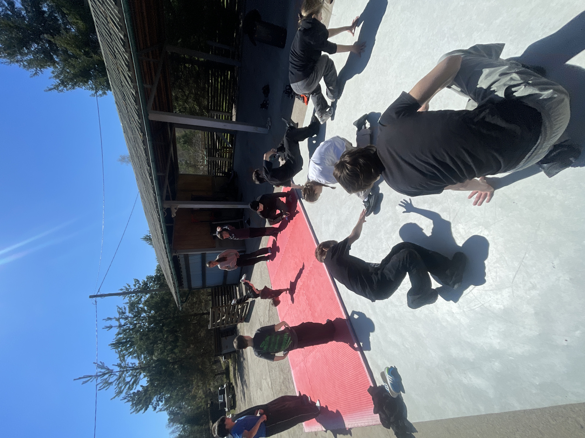 A group of approximately twelve people are performing exercises on a red mat and light-colored concrete surface. They are in various positions, some standing, some bending over, and some seemingly performing jumping or balancing movements. The scene appears to be outdoors, adjacent to a building with a covered structure. Beyond the building, trees are visible, and a clear blue sky is overhead. The location is identified as Daurskoe, Russia.