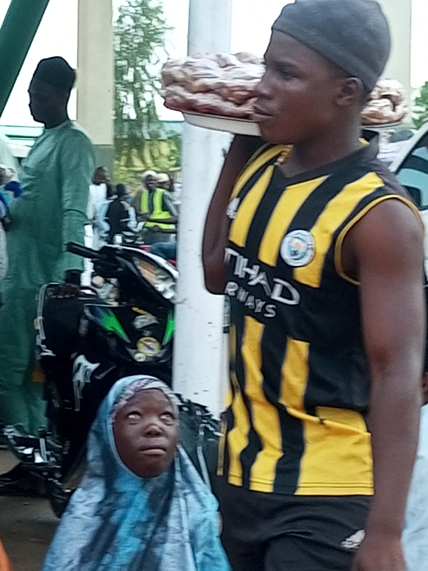 In Gombe, Nigeria, a young boy wearing a black and yellow striped jersey and a dark grey beanie stands facing right, carrying a tray of food. Beside him, a young girl in a light blue headscarf looks upwards with her eyes wide open. In the background, a person in a light green robe walks past a motorcycle. The scene appears to be outdoors, possibly in a market or public gathering area, with a light-colored pillar and trees visible in the background. The image captures a moment of daily life, with the boy appearing to be selling or distributing the food he carries, and the girl a passive observer in the scene.