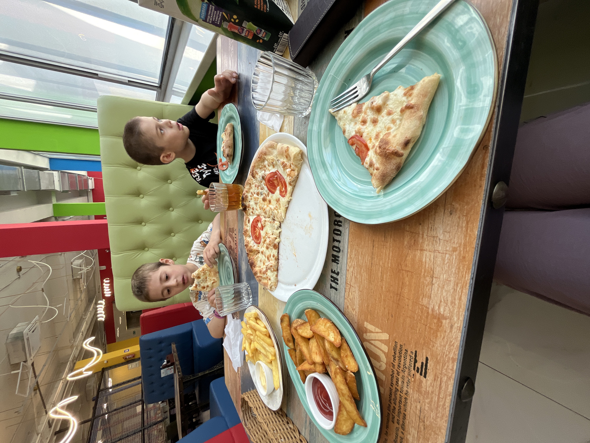 The image shows a table scene inside a restaurant, likely located in Kaspiysk, Russia. Two young boys are seated at the table, enjoying a meal. One boy is wearing a black t-shirt, while the other is wearing a white patterned t-shirt. In front of them are plates of pizza, french fries, potato wedges, and beverages. The food appears to be partially consumed. The table has a worn, wooden appearance with printed text, and the plates are a light green color. There are glass windows visible in the background, providing natural light. The restaurant features colorful interior design elements, including seating in various colors and decorative lighting fixtures.