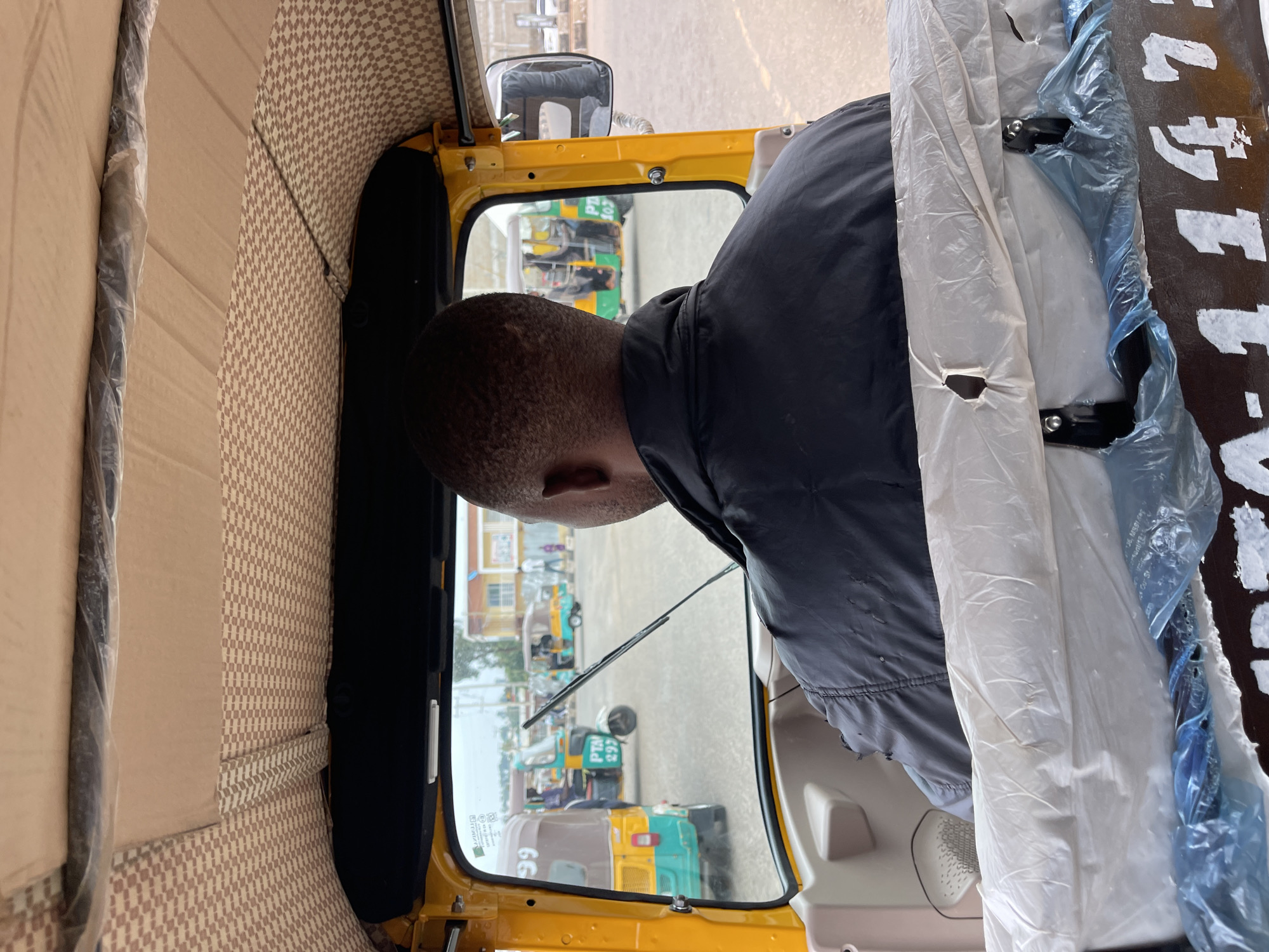 The image depicts the interior of a yellow auto-rickshaw in Jos, Nigeria. The view is from behind the driver, focusing on his head and shoulders. Outside the rickshaw, other similar vehicles are visible, indicating a transportation hub or a busy street. The environment is urban with buildings in the background. The auto-rickshaw's interior includes patterned upholstery. The vehicle seems new, as evidenced by the plastic covers on the seats. The driver, an African male with short hair, wears a dark jacket.