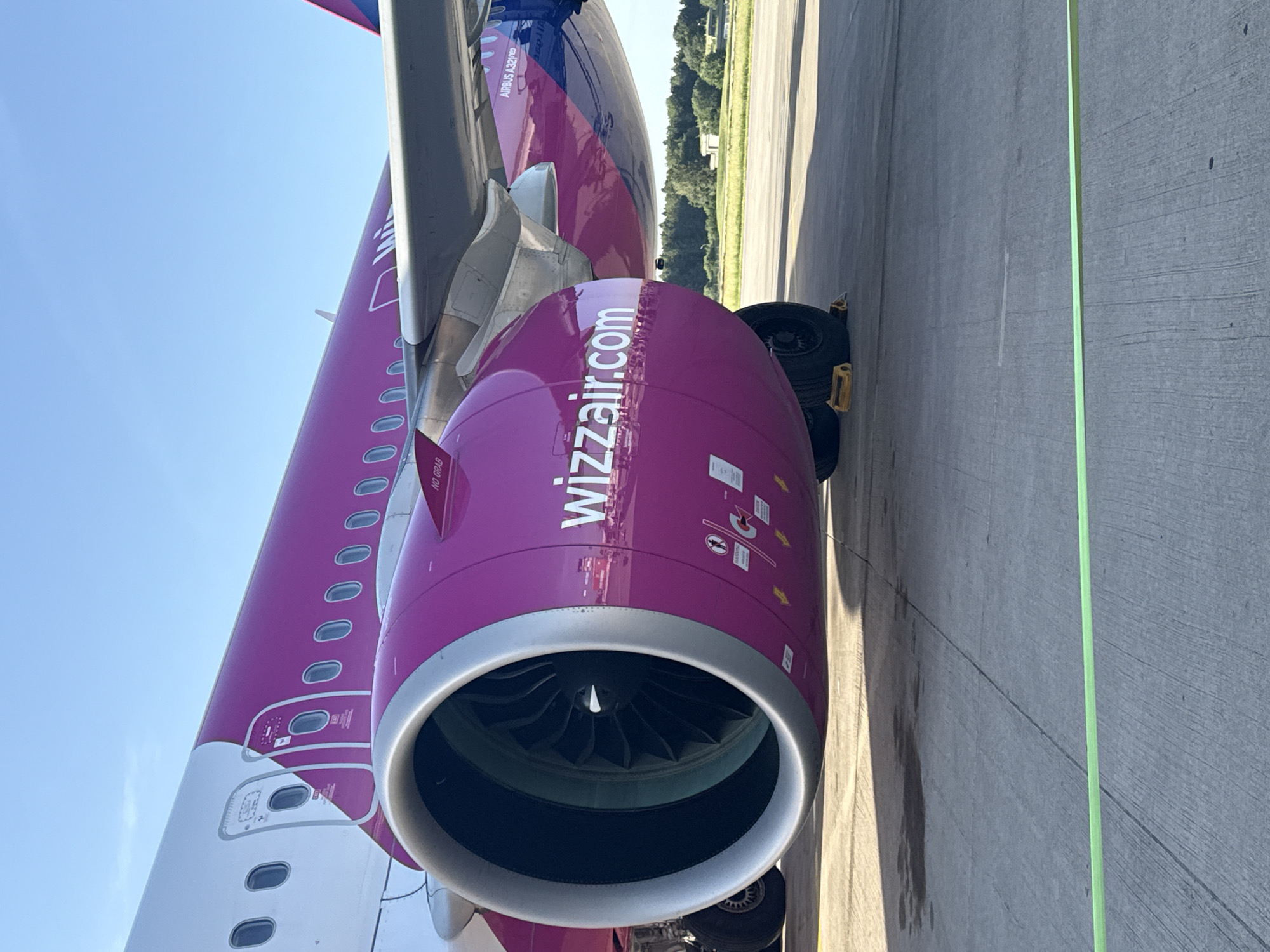 A detailed, close-up view of the engine and fuselage of a Wizz Air Airbus A321neo aircraft. The engine is prominently displayed with the "wizzair.com" logo. Part of the aircraft's fuselage, showing passenger windows, is visible. The scene takes place on an airport tarmac, identifiable by the concrete surface and painted lines. Trees in the distance imply a location near vegetation. The precise location of Künersberg, Germany, is not discernible from the image details alone.