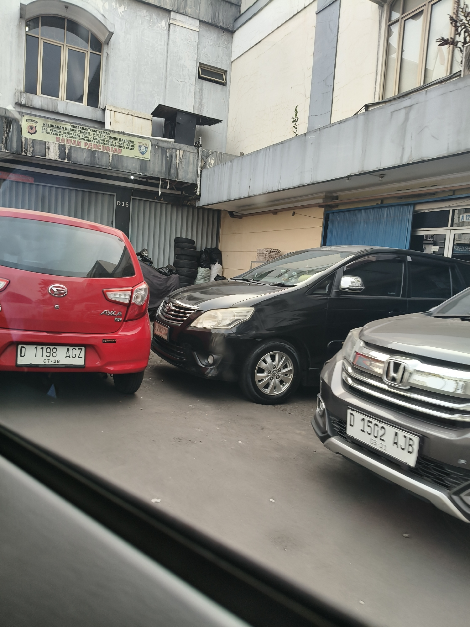 The image captures a street scene in Bandung City, Indonesia, focusing on several parked cars and the facade of a building. In the foreground, the rear of a red Daihatsu Ayla is visible, displaying a license plate that reads "D 1198 AGZ". To its right, a black Toyota Innova is parked, its front grille and headlights partially visible. Further to the right, the front of a gray Honda HR-V is seen, with the license plate "D 1502 AJB". Behind the cars, the building's exterior features a large arched window with multiple panes on the upper level and a banner with Indonesian text that appears to be an announcement or warning, mentioning "KEBON PISANG" and "SUMUR BANDUNG", suggesting a local area within the city. The building's architecture indicates it might be an official or commercial establishment. The overall environment appears to be an urban setting with street-level parking.