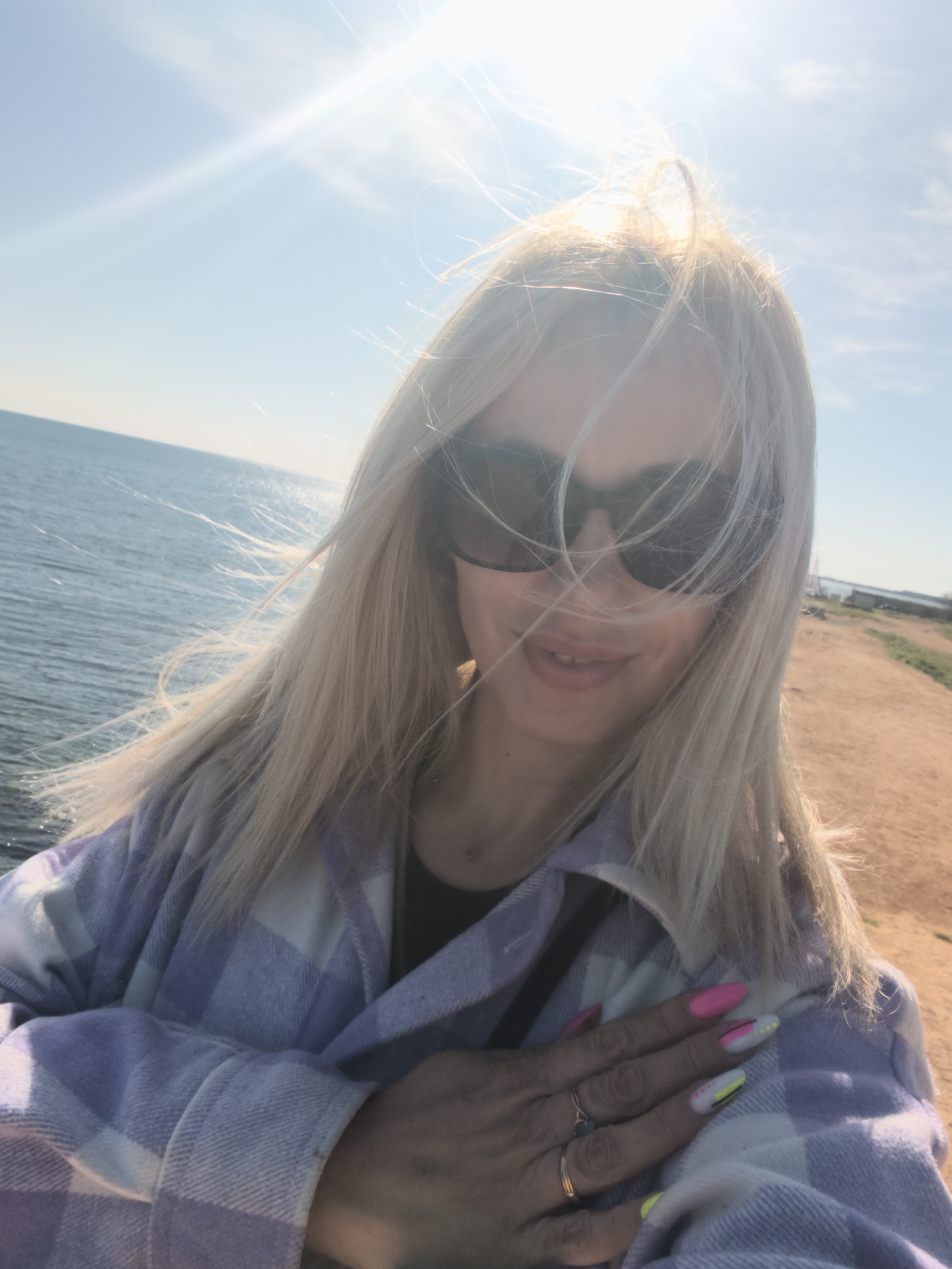 A bright, sunny selfie captures a young woman outdoors by the sea in Sevastopol, Ukraine. The woman, with long blonde hair, appears to be smiling faintly beneath dark sunglasses that cover her eyes. Her hair is windswept, blowing across her face and catching the sunlight. She is wearing a light purple and white plaid jacket over a black top. Her right hand, adorned with several rings on her fingers, rests on her chest, showcasing brightly manicured nails with pink, yellow, and white designs.
The background on the left features a vast expanse of blue sea or ocean, with sunlight reflecting off the water's surface. On the right, a sandy or dirt path runs along the coastline, suggesting a natural embankment or beach. The sky is clear blue, and the sun, positioned in the upper left, casts a strong, bright flare, indicating a very sunny day, likely midday or afternoon. The overall scene suggests a casual, breezy moment by the water. There is no visible text in the image.