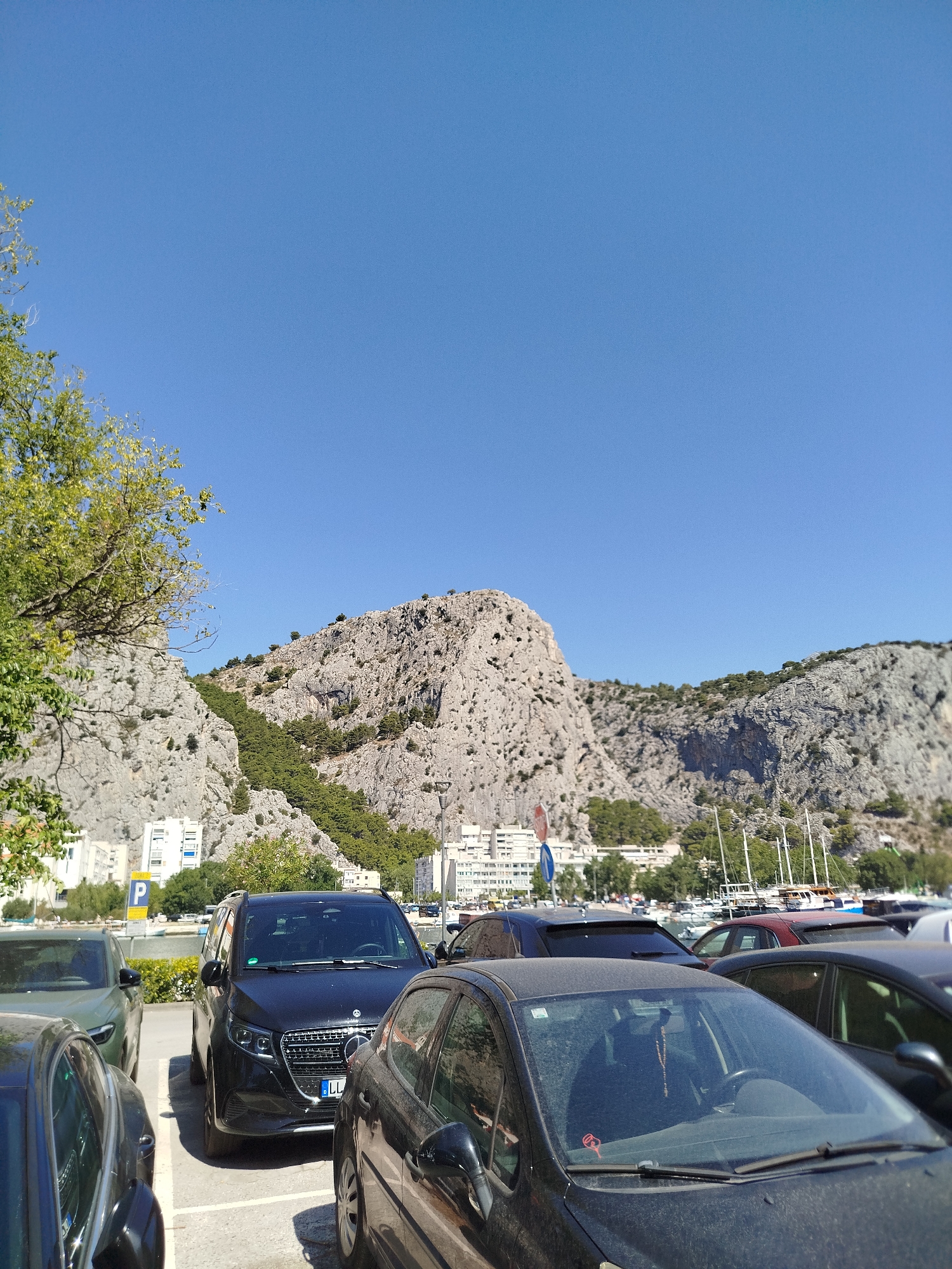 The image depicts a parking lot in Omiš, Croatia, with a prominent rocky mountain range in the background. Several cars are parked, including a black Mercedes-Benz van in the center, a dark-colored sedan to its left, and another dark sedan partially visible in the foreground. The mountain, covered with sparse vegetation and trees, rises steeply behind a row of white buildings. In the distance, sailboats are docked along the waterfront, their masts creating a distinct silhouette against the sky. The sky is clear and blue. A yellow parking sign with a blue 'P' is visible to the left of the Mercedes van. The overall scene suggests a sunny day in a coastal town.