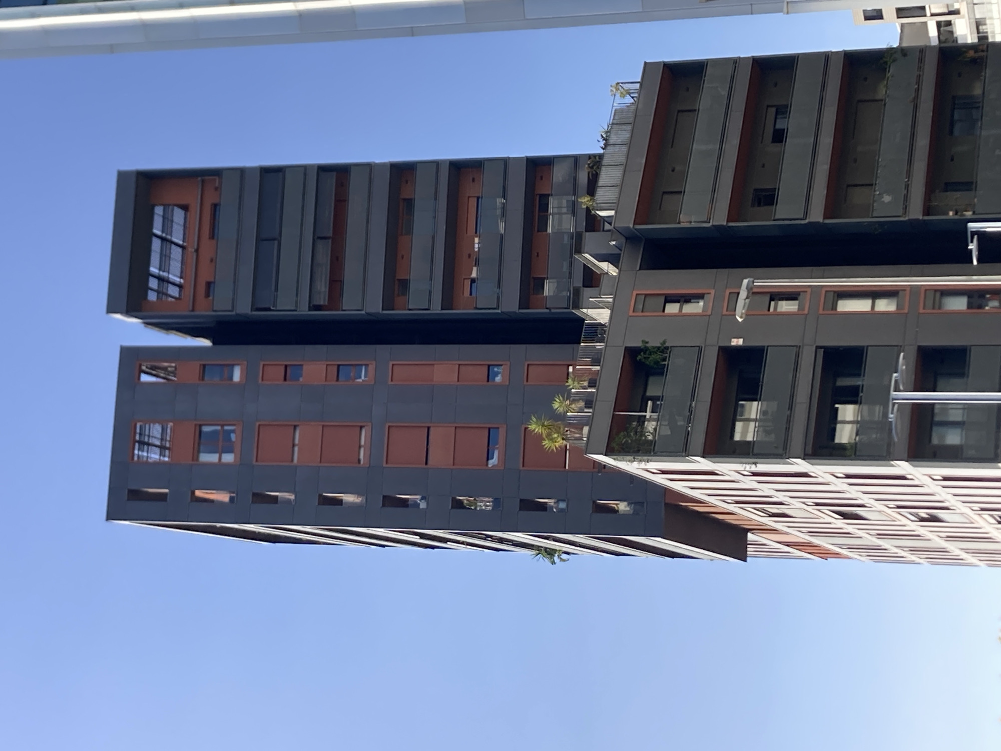 This is a photograph of modern, high-rise residential buildings against a clear blue sky. The buildings feature a distinctive architectural style with alternating sections of dark gray, textured paneling and warm, reddish-brown rectangular panels. Many windows are visible, some appearing as dark rectangles and others reflecting the sky. The buildings have sharp, angular designs with cantilevered sections. Vegetation, including what appears to be palm fronds, is visible on balconies and rooftops, suggesting a warm climate. The photograph is taken from a low angle, looking upwards, which emphasizes the height and scale of the structures. The sunlight indicates it is daytime, likely a sunny day. The overall impression is one of contemporary urban architecture in a sunny location. No people or specific events are depicted.