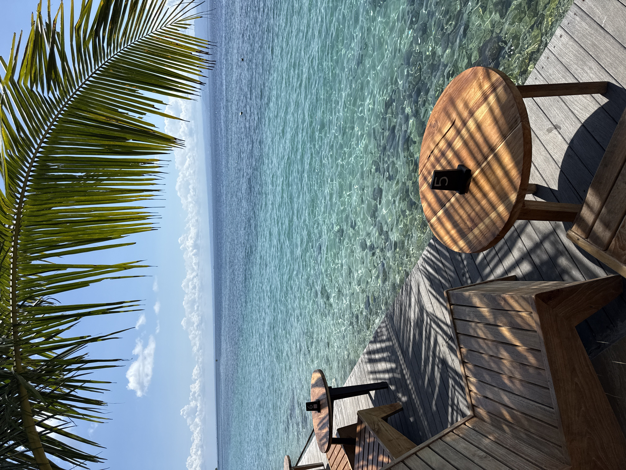 A serene, outdoor scene presents an elevated wooden deck in Nusa Penida, Indonesia, juxtaposed against the tranquil turquoise waters. The focal point of the setting is a small, round wooden table with the number "5" affixed on its surface, coupled with complementary wooden seating. To the left, a verdant palm frond partially frames the scene. The clear waters reveal the underwater terrain, hinting at the proximity to the shore. The sky above exhibits a combination of clear blue patches interspersed with cumulus clouds. The photograph captures an atmosphere of relaxation and tropical ambiance.