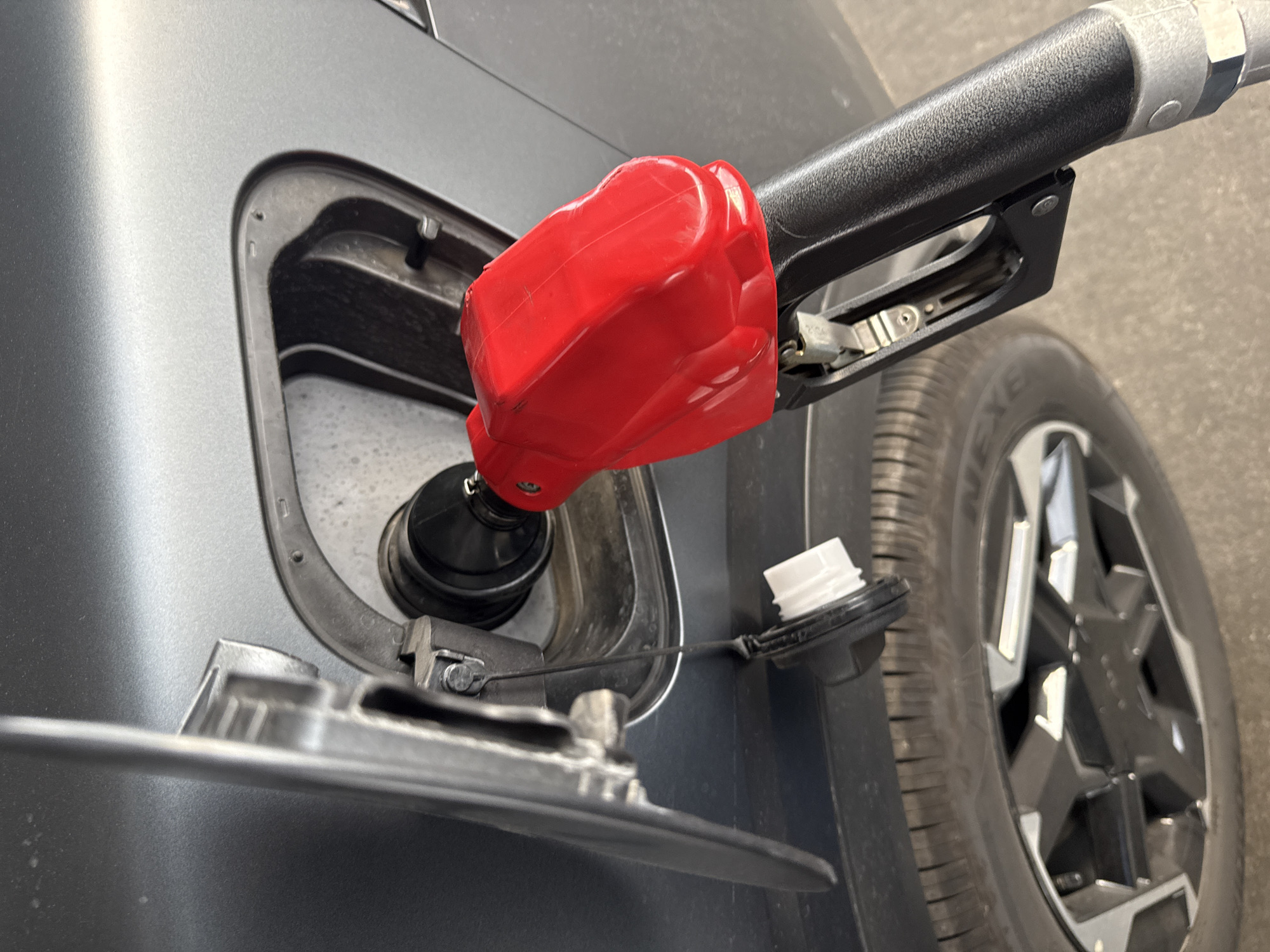 A gray car is being refueled at a gas station. A red gas nozzle is inserted into the car's fuel tank. The fuel tank door is open, and the cap is attached to the car by a tether. A tire and the pavement of the gas station are visible. The specific location in Puebla, Mexico, cannot be determined from the given image.
