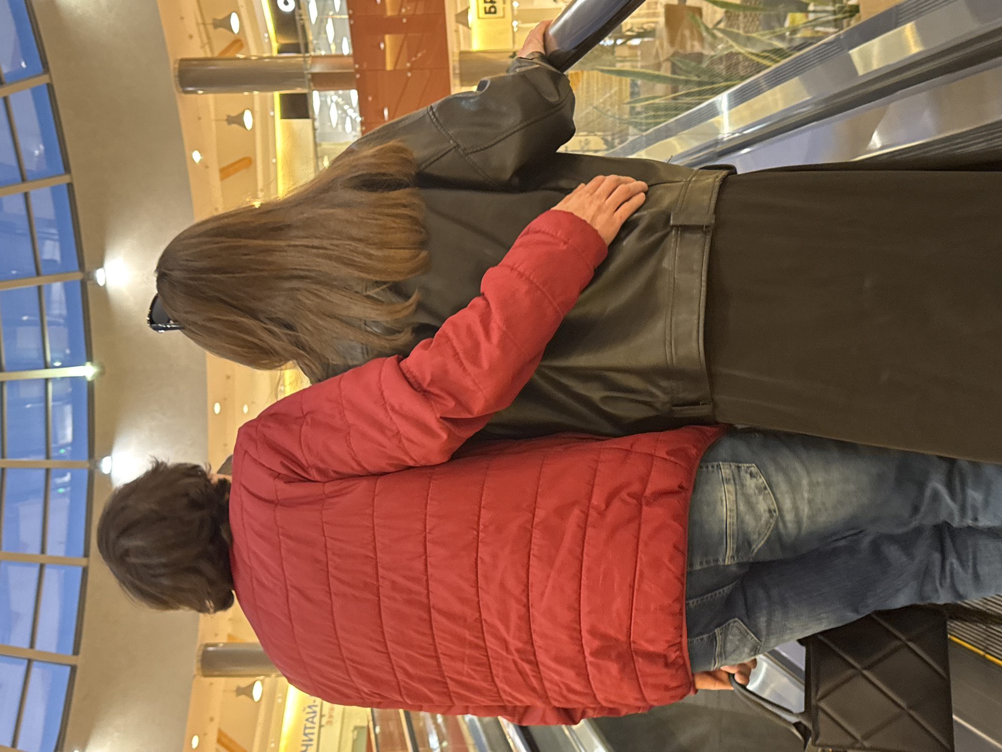 A rear view shows two people on an escalator, likely in a shopping mall based on the blurred background. The person in front has long, brown hair and wears a black leather jacket, black skirt or dress. The person behind wears a red puffer jacket and jeans. Their arm is draped across the front person's back. The escalator is metallic with visible handrails. The location appears to be Perm, Russia based on a sign that includes Russian text.