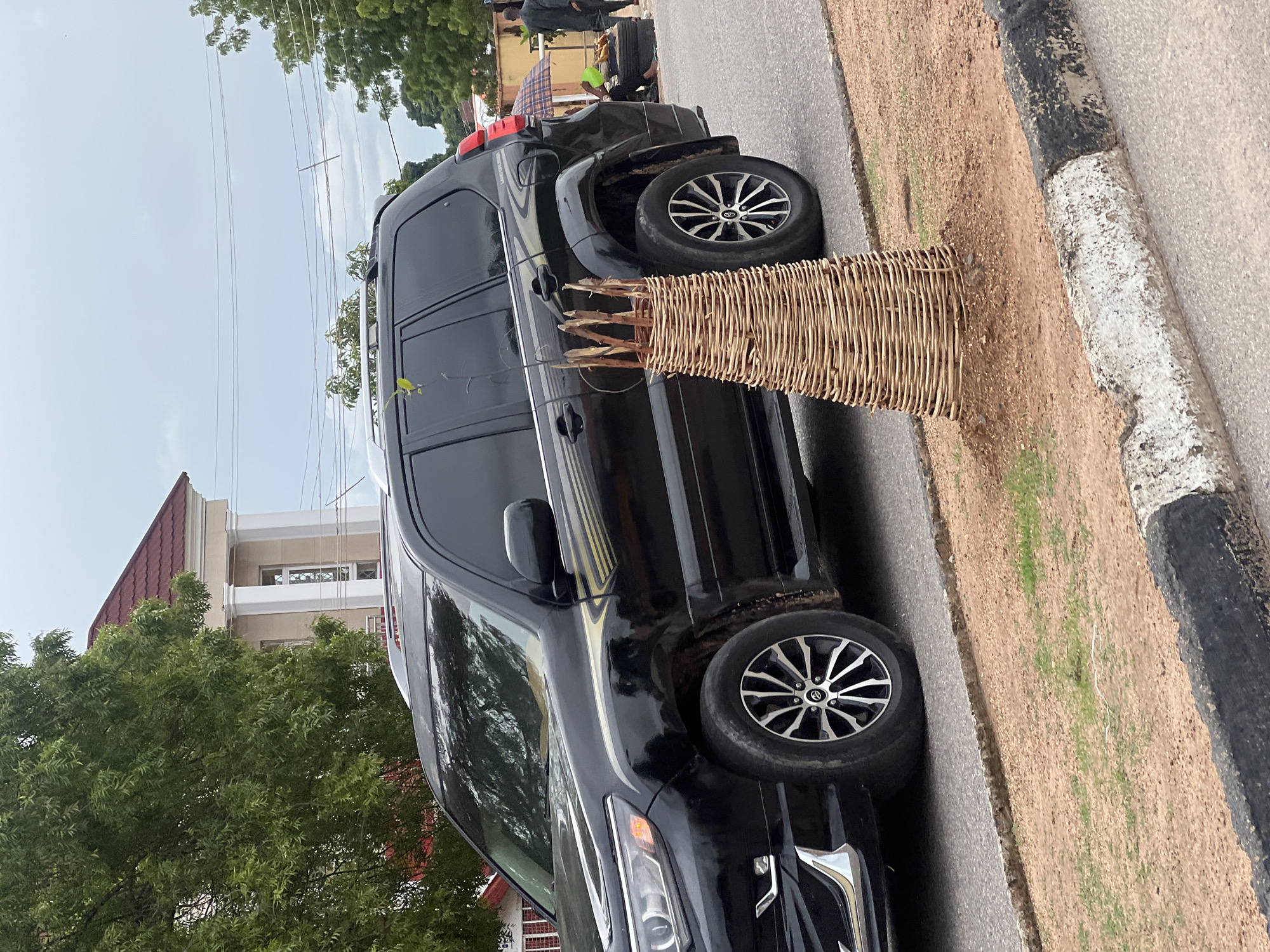 A black SUV is parked on the side of a road in Jalingo, Nigeria. In front of the SUV, a young tree is protected by a woven basket-like structure. The tree has a thin, brown stem with a few green leaves. The woven structure is conical in shape and made of light-colored reeds. The SUV is parked on asphalt, with a dirt and grass area to its right. In the background, there is a large building with a red tiled roof and white columns. A few trees are visible on the left side of the image. Further down the road, two people are visible under a large umbrella, with some tires and goods arranged on the ground. The sky is overcast.