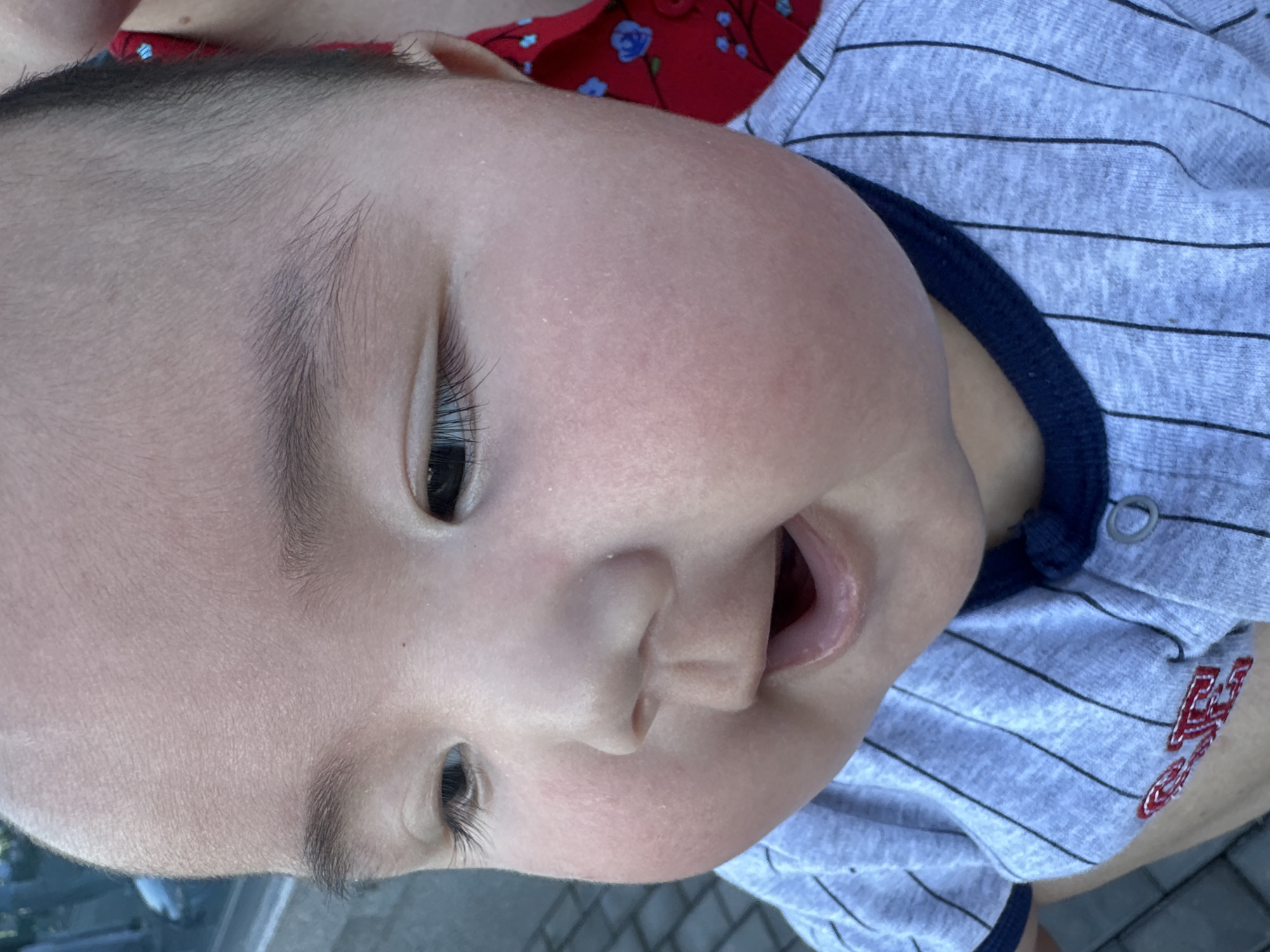 This media file is a close-up, vertical, eye-level shot of a young child's face, captured outdoors. The child is wearing a grey and black striped t-shirt with a dark blue collar. A portion of a red garment with a floral pattern is visible behind the child's head.
The child's face is the central focus, with their eyes, nose, and mouth prominently displayed. The skin appears fair and smooth, with a slight rosy hue on the cheeks. The child's left eye is open and looking slightly to the side, while the right eye is partially closed. The child's mouth is open, revealing their tongue and what appear to be developing teeth. The child's eyebrows are thin and dark.
The background is blurred but suggests an outdoor setting with paved ground, possibly a sidewalk or patio, visible at the bottom of the frame. The lighting appears to be natural daylight, suggesting the photo was taken during the day. There is no discernible text in the image. The overall impression is an candid, intimate portrait of a young child.