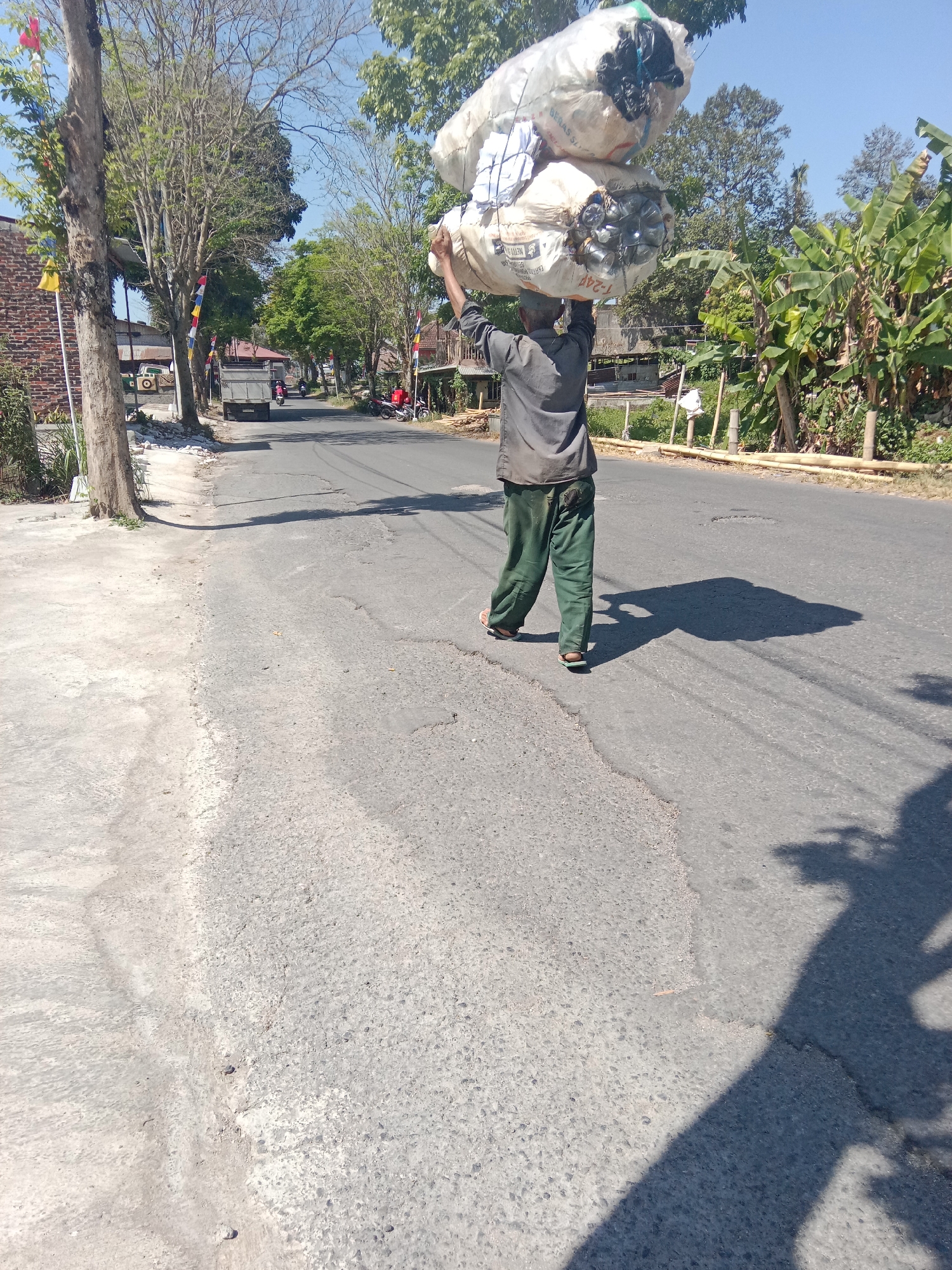 In the city of Parakan Kauman, Indonesia, a person is walking along a paved road, carrying two large sacks of what appears to be recyclable materials balanced on their head. The sacks are white and filled with various items, including plastic bottles and possibly other discarded materials. The individual is wearing a dark grey shirt and loose green pants, with sandals on their feet. They are walking away from the camera, casting a long shadow on the road. The environment is outdoors, with a clear blue sky above. Along the side of the road, there are trees, some with dry branches, and vegetation, including banana plants. In the background, there are buildings and a few motorbikes and a truck, suggesting a residential or semi-rural area. The road itself shows signs of wear and tear with visible cracks and patches. The image captures a moment of everyday life, highlighting the labor involved in collecting and transporting such materials.