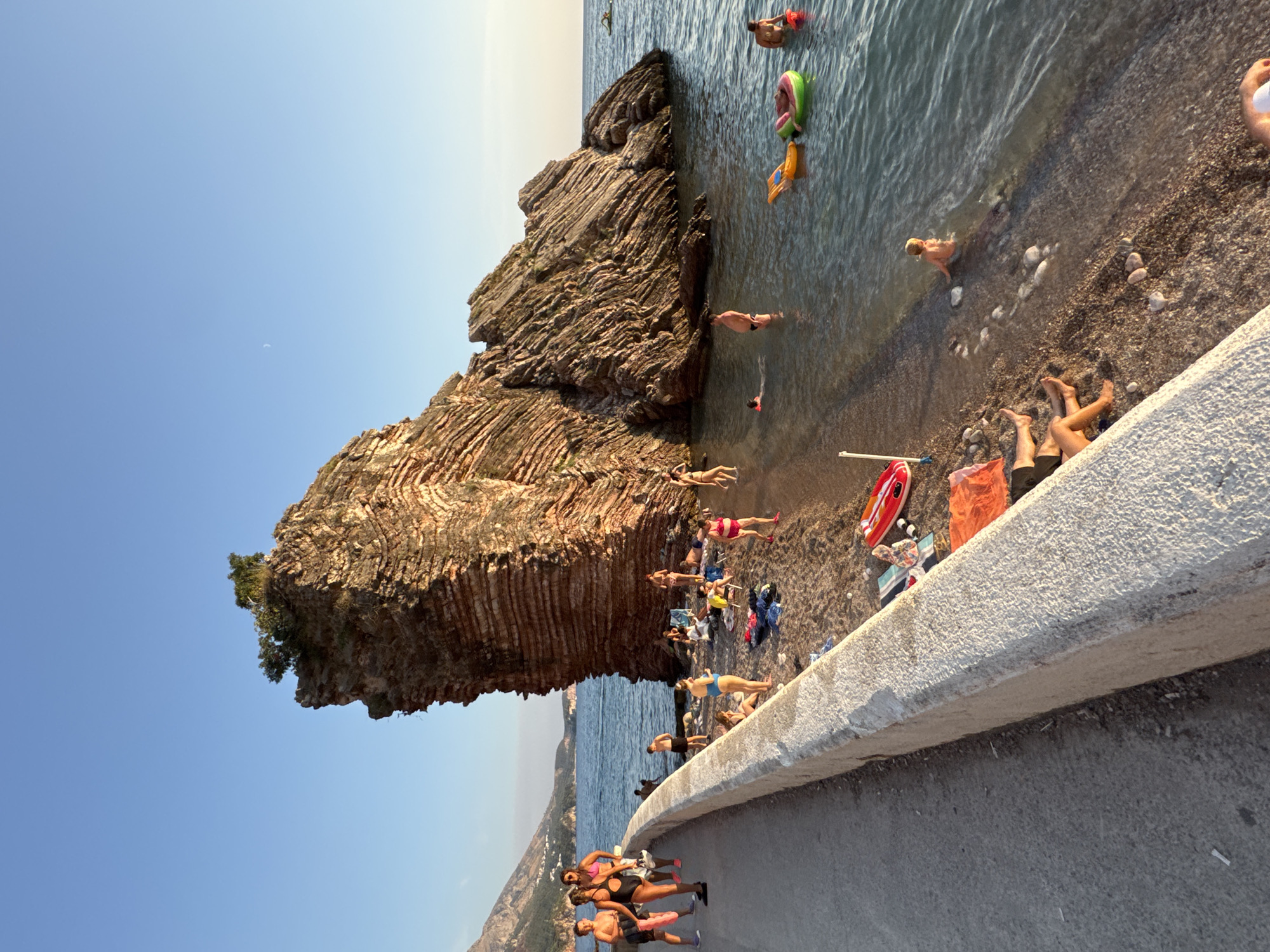 The image depicts a beach scene in Kamenovo, Montenegro, dominated by a large, layered rock formation jutting out of the sea. Several people are present on the pebble beach and in the shallow water. On the left side of the image, a paved walkway runs along the beach, bordered by a low concrete wall. A group of people, including children, are walking along this path. On the beach itself, individuals are sunbathing on towels, sitting in chairs, and wading in the water. Some are engaged in swimming or playing in the sea. Inflatable toys, such as a yellow ring and a green ring, are visible in the water. The sea is calm and clear, reflecting the blue sky. The sun appears to be setting or rising, casting a warm light on the scene. In the background, mountains are visible on the left, and the sea extends to the horizon. A faint crescent moon is visible in the clear blue sky.