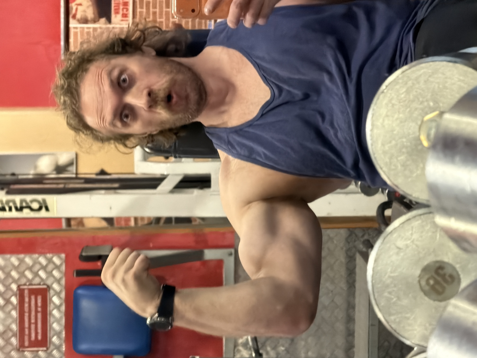 A man with curly brown hair and a mustache is taking a selfie in a gym. He is wearing a navy blue tank top and a black wristwatch on his left wrist. He is holding a dumbbell in his right hand and flexing his bicep. The dumbbell weighs 38 kilograms. The background includes gym equipment, a red wall, and a poster. The location cannot be determined from the information provided.