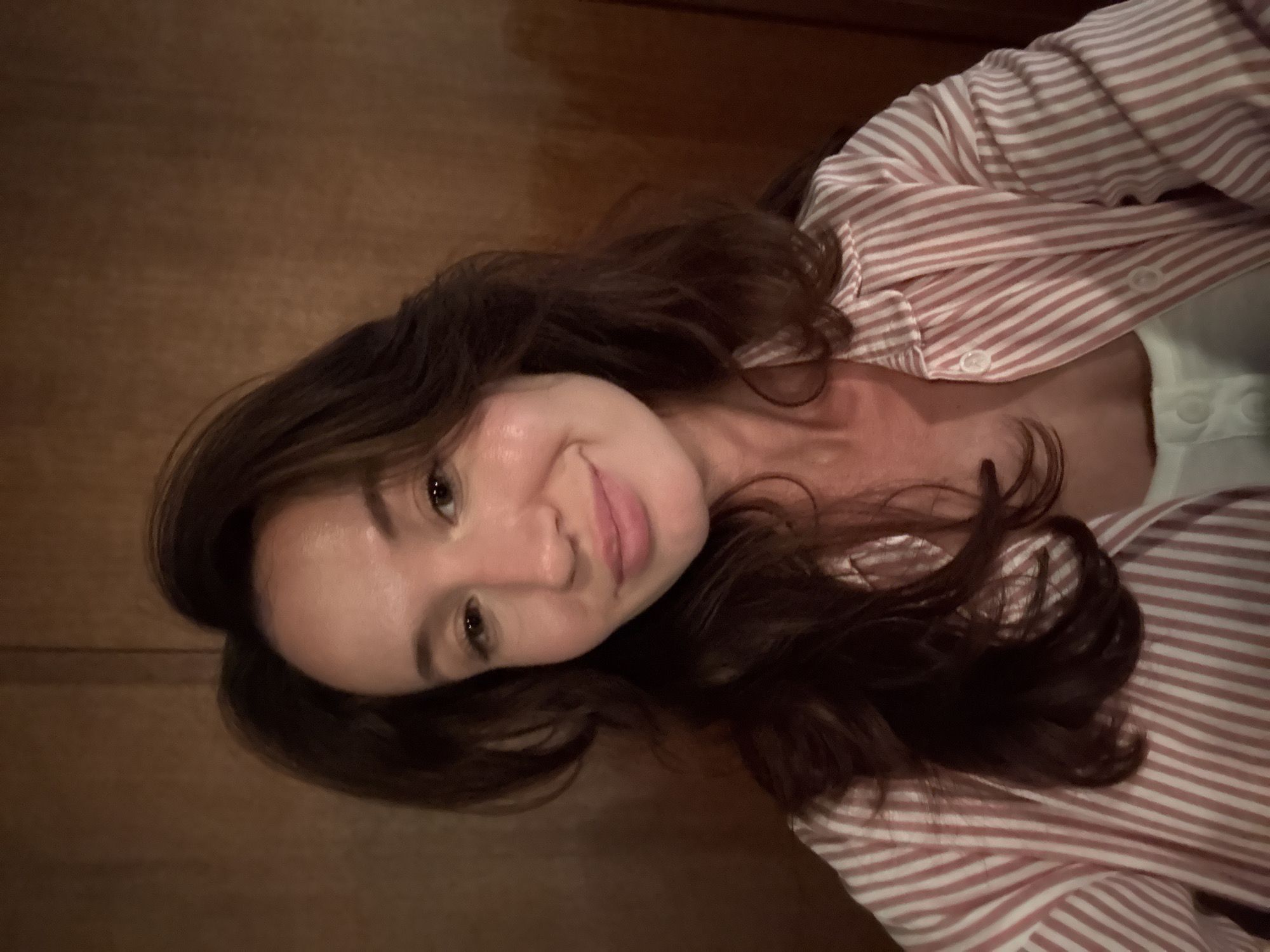 A woman with long, dark brown hair and fair skin is photographed in a close-up, upper body shot. She is wearing a pink and white striped button-up shirt with the collar open. Her head is tilted slightly to her left, and she is looking directly at the camera with a gentle smile. Her skin appears dewy and possibly has a subtle glow. The background is a warm, dark brown, suggesting a wooden wall or paneling. The lighting is soft, highlighting her features and creating a warm ambiance. There is no visible indication of the city of Tel Aviv, Israel.