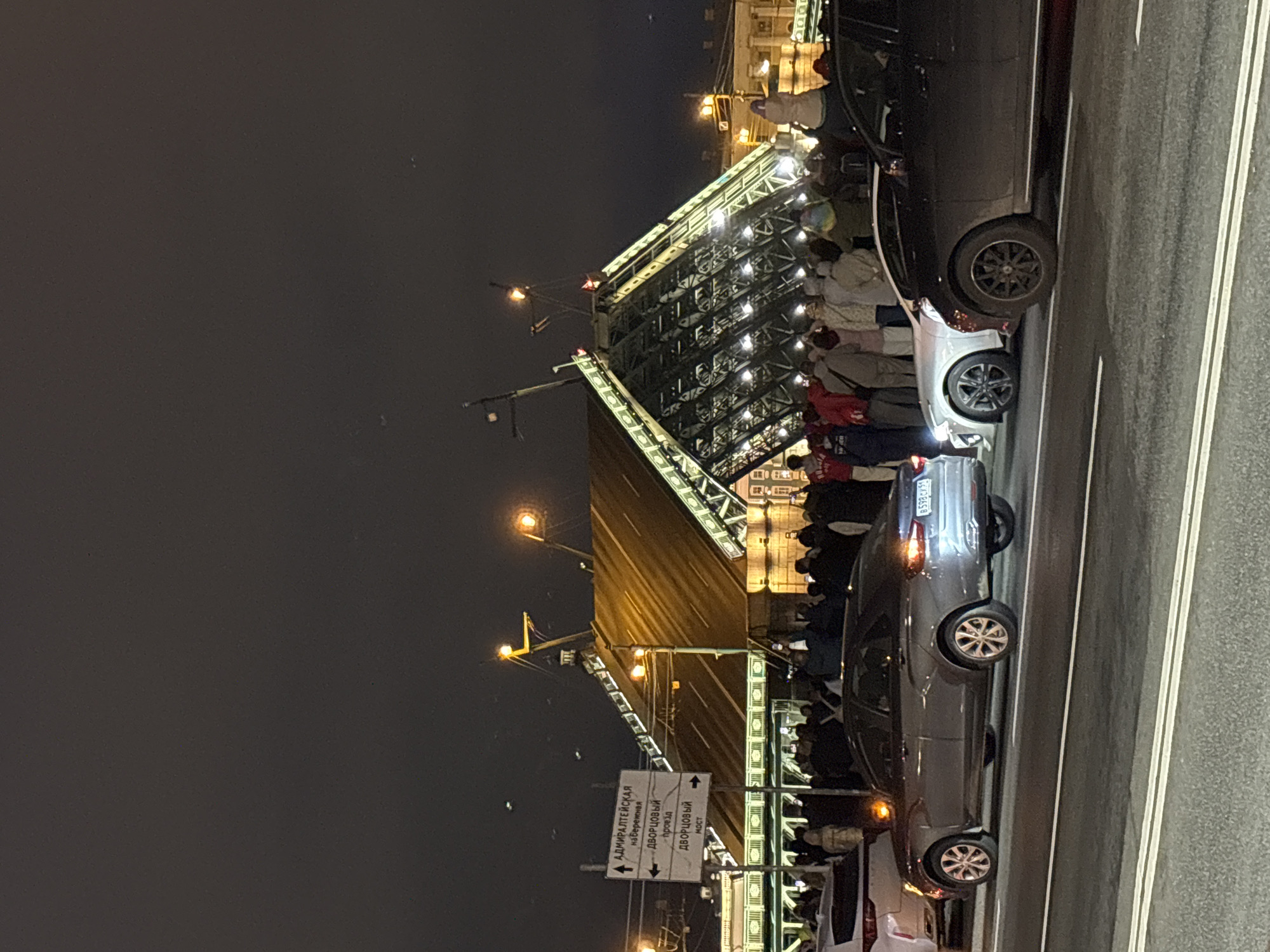 At night, a large crowd watches the Palace Bridge (Dvortsovyi Most) rise in Saint Petersburg, Russia. Cars are parked in the foreground along a road with white lane markings. Streetlights and bridge lights illuminate the scene. A street sign directs traffic to the Admiralty Embankment and Palace Bridge.