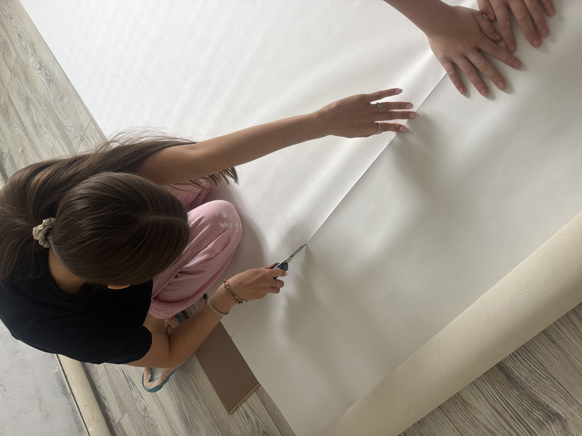 A high-angle shot depicts a woman cutting a large sheet of white paper or wallpaper with a utility knife. She is wearing a black T-shirt, pink pants, and blue slippers. Her hair is dark and pulled back in a ponytail. A child's hands are visible on the top-right of the image, holding the paper. A roll of paper is located on the bottom-right of the image. The floor is a light, wood-like surface. A piece of cardboard is near the woman's legs on the left-side. The photo was not taken in Markovo, Russia, as there is no information to confirm this.
