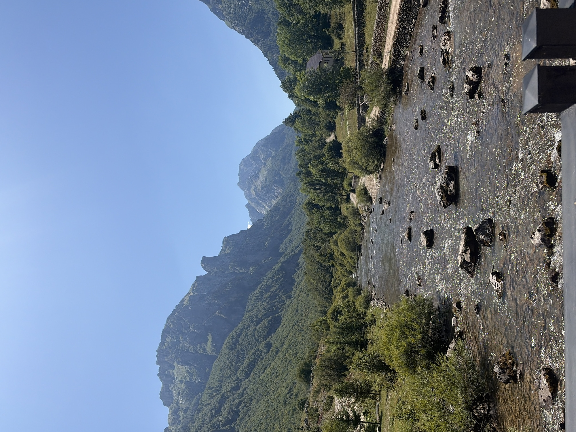 The image shows a wide river flowing through a valley surrounded by mountains. The water is clear and shallow in places, with rocks visible on the riverbed. The riverbanks are covered with lush green vegetation, including trees and bushes. In the distance, rugged mountains rise sharply against a clear blue sky. A small, light-colored house with a dark roof is nestled among the trees on the right bank of the river. A dirt path runs alongside the riverbank, bordered by stone walls. The overall impression is one of a serene natural landscape. The location is likely the city of Vusanje, Montenegro, known for its mountainous terrain and clear rivers. There are no people visible in the image.