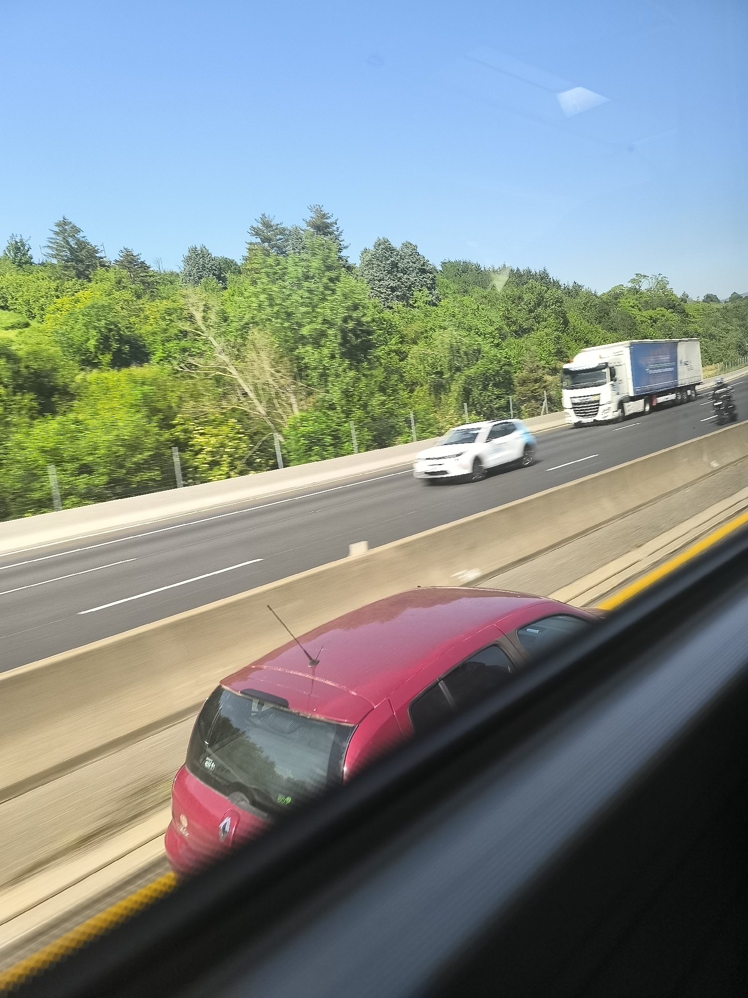 The image shows a multi-lane highway near L'Horme, France. Several vehicles are visible, including a red hatchback, a white car, a semi-truck, and a motorcycle. The highway is bordered by a concrete barrier and lush green trees. The sky is clear and blue. The perspective suggests the image was taken from a moving vehicle on the highway.