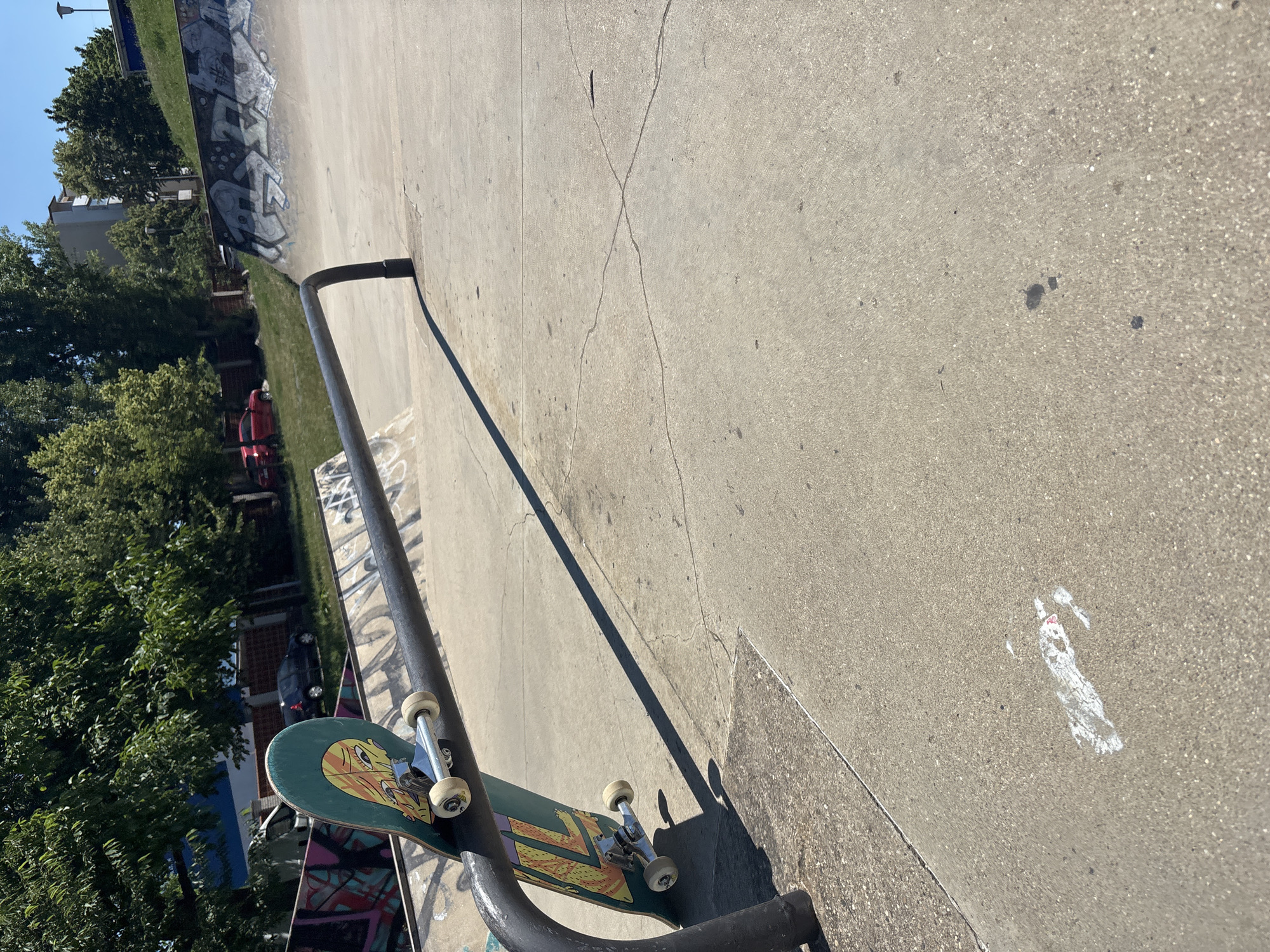The image shows a skateboard leaning against a metal rail in a skatepark. The skateboard has a green deck with a cartoonish face graphic. The wheels and trucks are silver. The skatepark appears to be made of concrete and has graffiti on some of its surfaces. In the background, there are trees and buildings, suggesting an urban environment. The sun is shining, casting a long shadow from the rail across the concrete.