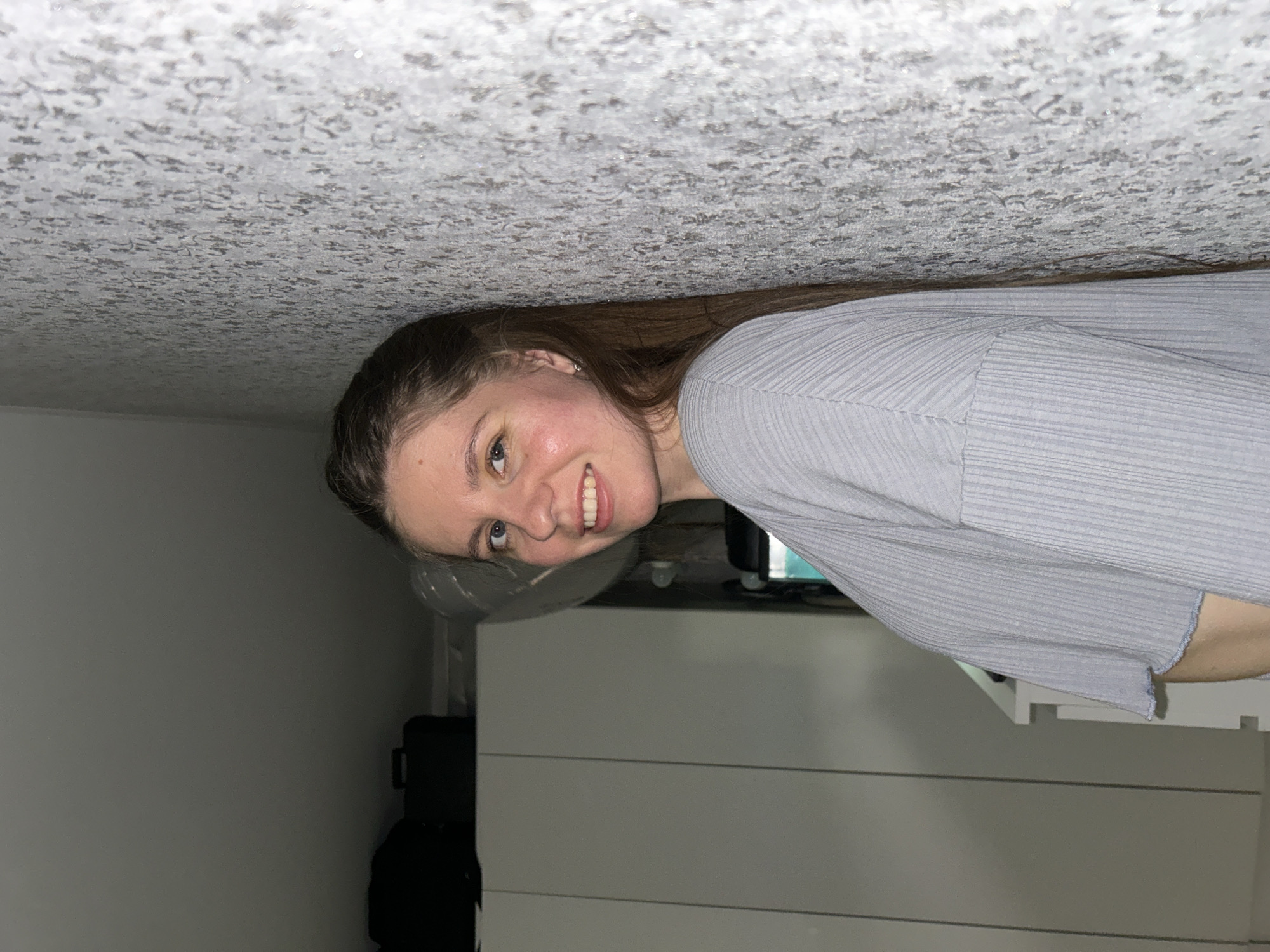 A woman with light brown hair is visible. Her face is partially obstructed by what appears to be a ceiling covered in a textured, possibly floral-patterned surface. She is wearing a gray, ribbed shirt. Behind her, there is a white dresser or cabinet with horizontal lines. A black bag is visible on the left. There is no information in the image to determine if it was taken in Klin, Russia.