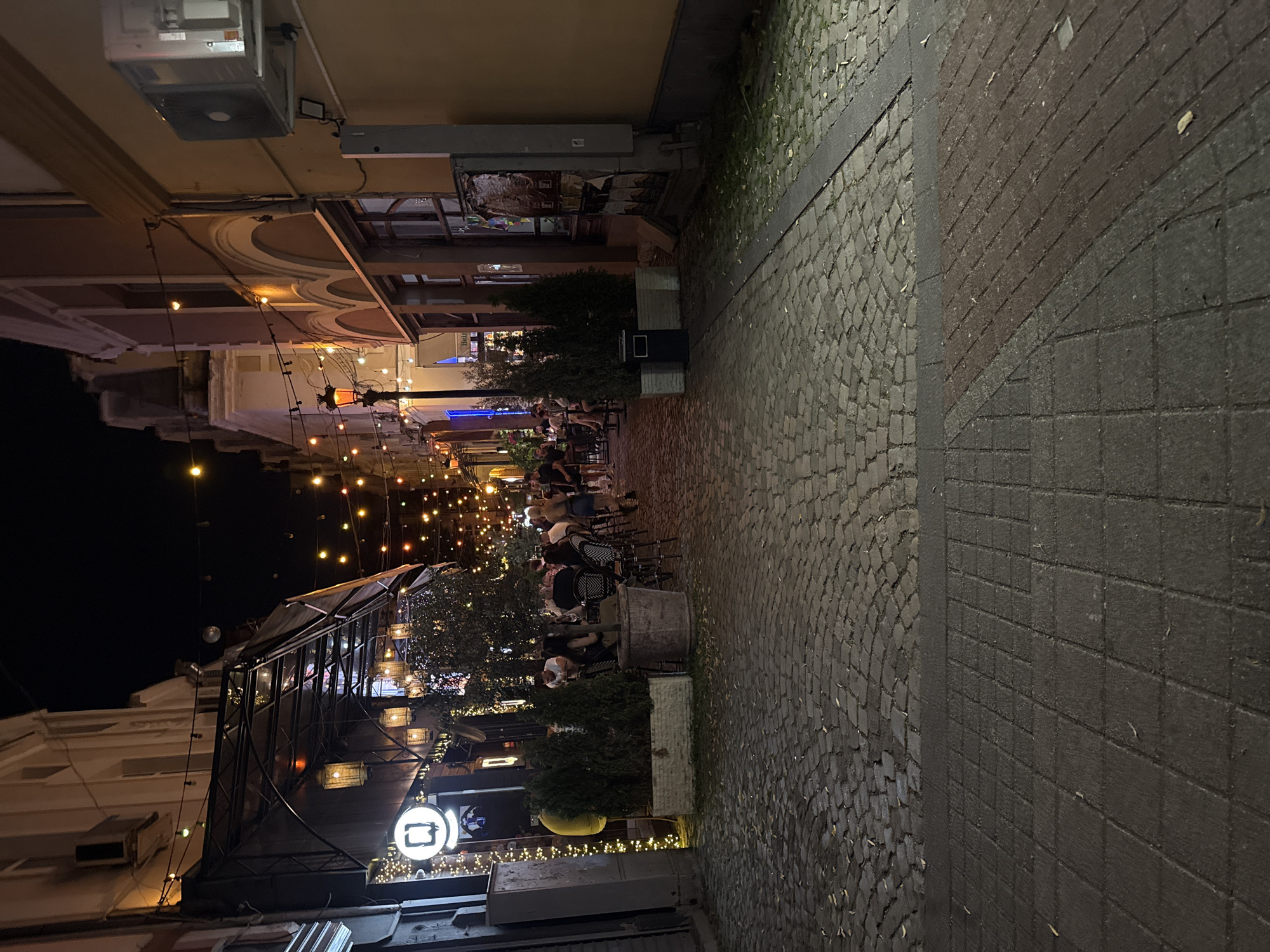 The image depicts a cobblestone street at night in the Kapana district of Plovdiv, Bulgaria. String lights are draped across the street, illuminating an outdoor dining area. Several people are seated at tables outside a restaurant, some engaged in conversation, while others appear to be waiting or eating. The architecture of the buildings lining the street is visible, with some featuring arched windows and balconies. The overall atmosphere is lively and social, indicative of a popular nightlife spot.