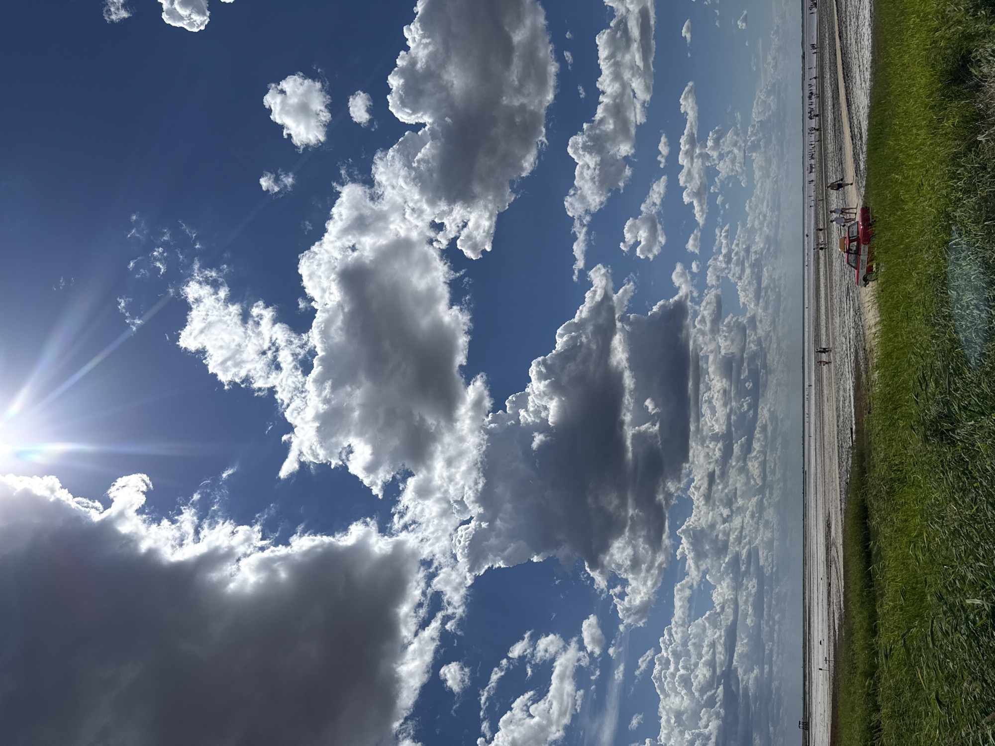A bright, sunny day is depicted with dramatic, fluffy white and grey clouds filling a vibrant blue sky. The sun is positioned in the upper left, casting bright rays. The scene appears to be the coastal area of Bursol, Russia, possibly near a salt lake or a shallow body of water. In the foreground, lush green grass sways. Further back, a vast, flat expanse stretches towards the horizon, dotted with many small figures of people. A red vehicle with a yellow roof is parked on a dirt path in the middle ground, with a few people standing nearby. The overall impression is one of a lively outdoor scene on a clear summer day.