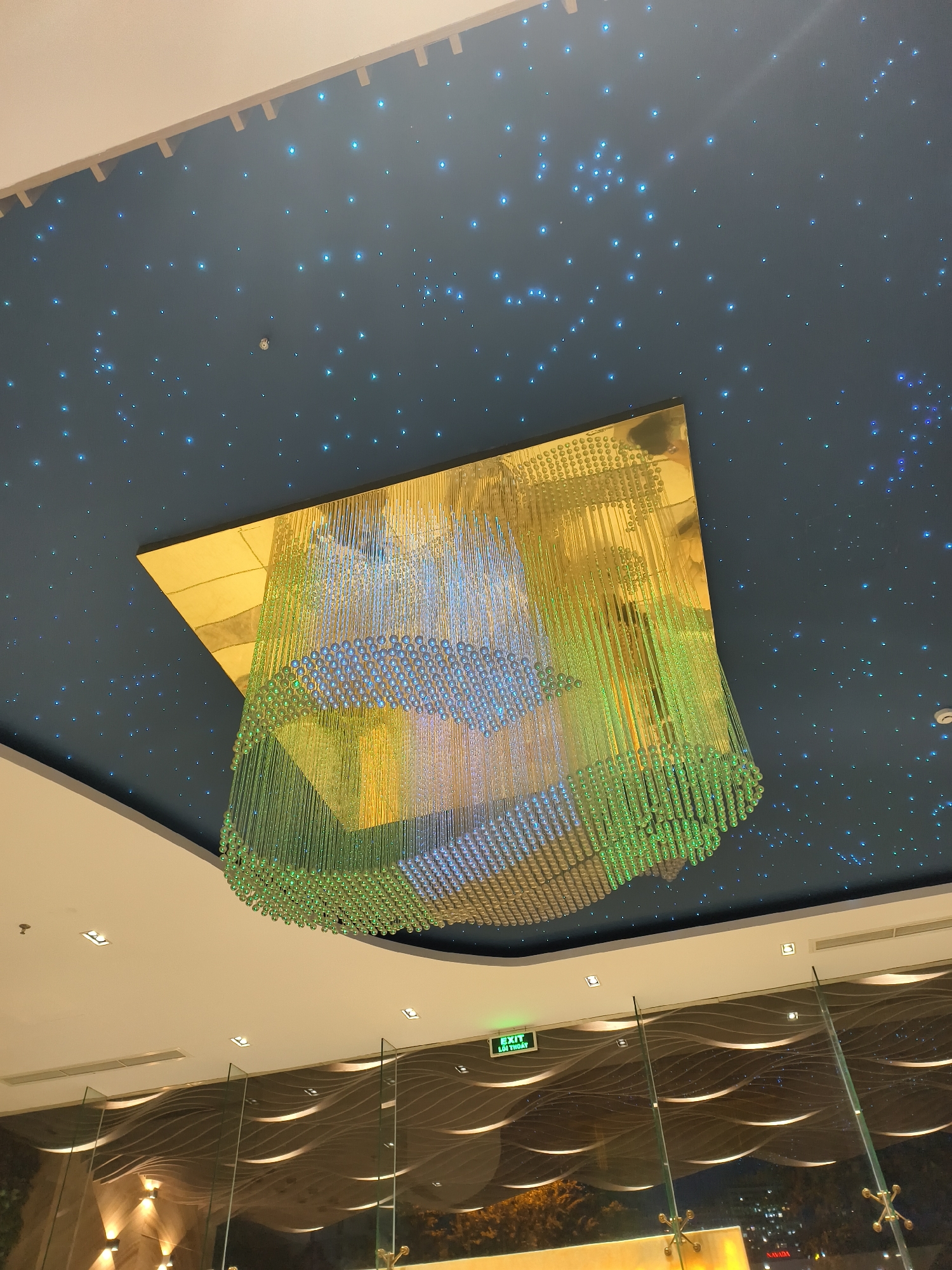 The image shows a close-up, low-angle view of an indoor ceiling and chandelier. The ceiling is dark blue and speckled with numerous small, bright blue lights, resembling a starry night sky. A large, modern chandelier hangs from a square, gold-colored fixture in the center of the frame. The chandelier itself is composed of many strands of clear, spherical beads that transition in color from blue at the top to green at the bottom. The lighting in the scene is dim, with the exception of the small ceiling lights and the illuminated chandelier. Below the ceiling, a white soffit is visible, and beneath that, a glass partition with a wave-like pattern and some recessed square lights. An illuminated green "EXIT" sign is also visible on the white soffit. The overall impression is one of a modern, upscale interior space.
