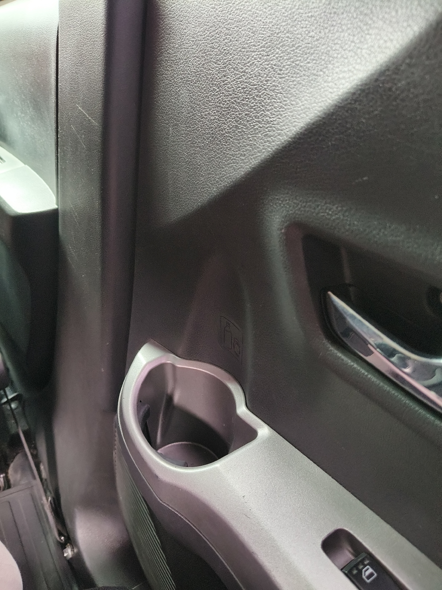 The image shows a close-up of the interior of a car, likely a door panel. The dominant colors are gray and black, with textured surfaces typical of automotive plastic. A cup holder is visible in the lower center of the frame, with its opening facing the viewer. Above and to the left of the cup holder, there is a small icon embossed into the plastic that appears to be a bottle with a line through it, possibly indicating a "no drinking bottle" symbol. Further to the left, part of a silver door handle or trim piece is visible. In the lower right corner, a car window control button with an icon of a window is present. The overall impression is that of a functional, utilitarian car interior. There are no people or any indications of the city of Bandung, Indonesia, in the image.
