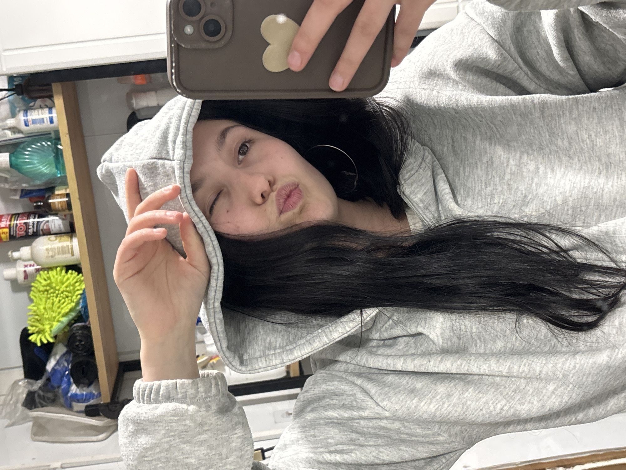 A young woman with long, dark hair and wearing a light gray hooded sweatshirt is taking a selfie in a mirror. She is winking one eye and puckering her lips, holding a smartphone with a brown case and a heart-shaped attachment above her forehead. The background shows a cabinet with various items, including bottles and a green sponge. The location cannot be determined to be Фоки, Russia from the image alone.