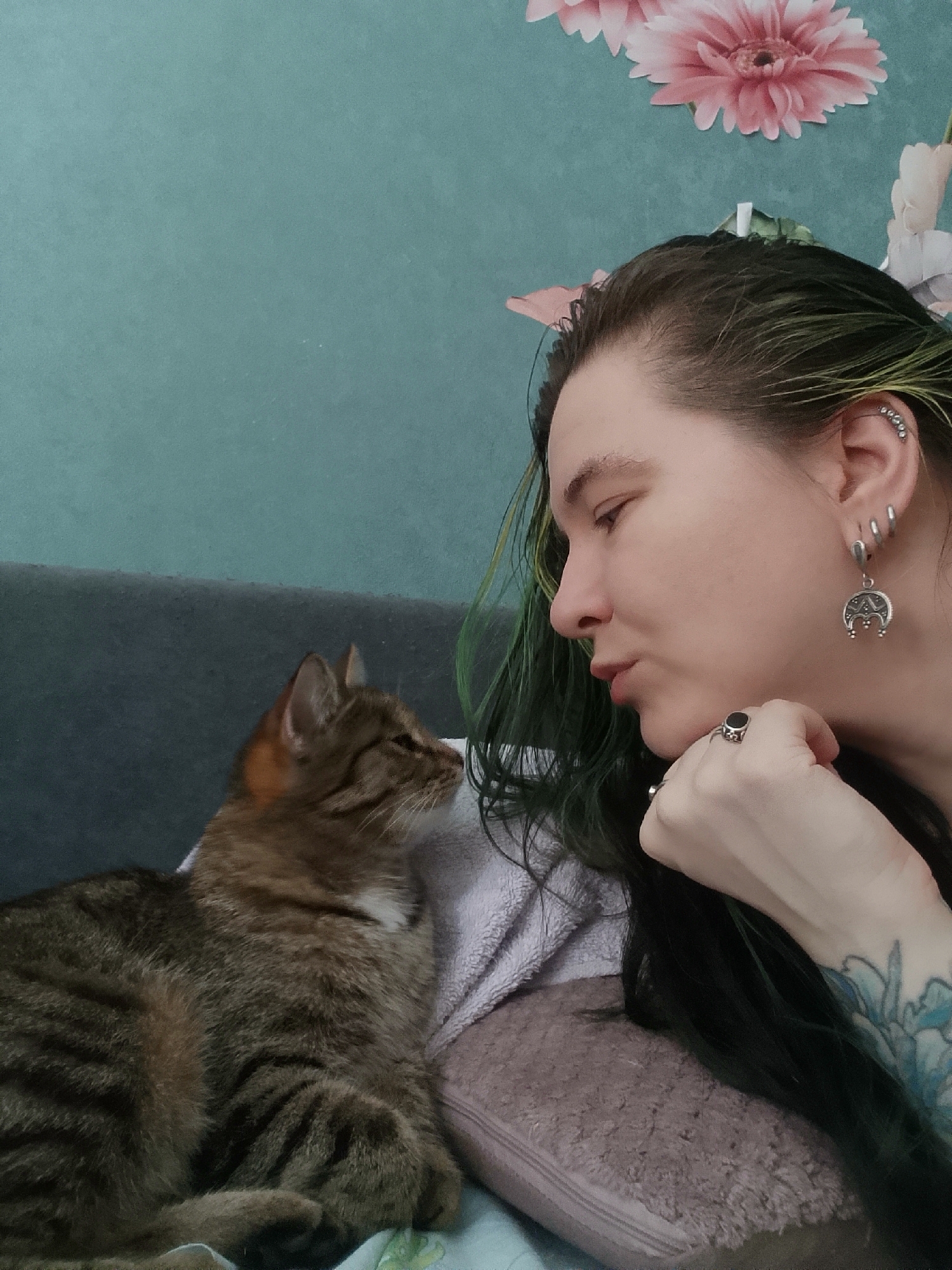 The image shows a woman and a cat. The woman is lying down, facing left, with her head resting on her hand. She has green-streaked hair, multiple piercings in her left ear, and rings on her fingers. A tabby cat is lying next to her, also facing left. The background consists of a gray couch, a teal wall with floral decorations, and a lavender towel. The city of Chaikovsky, Russia is not identifiable from this image.