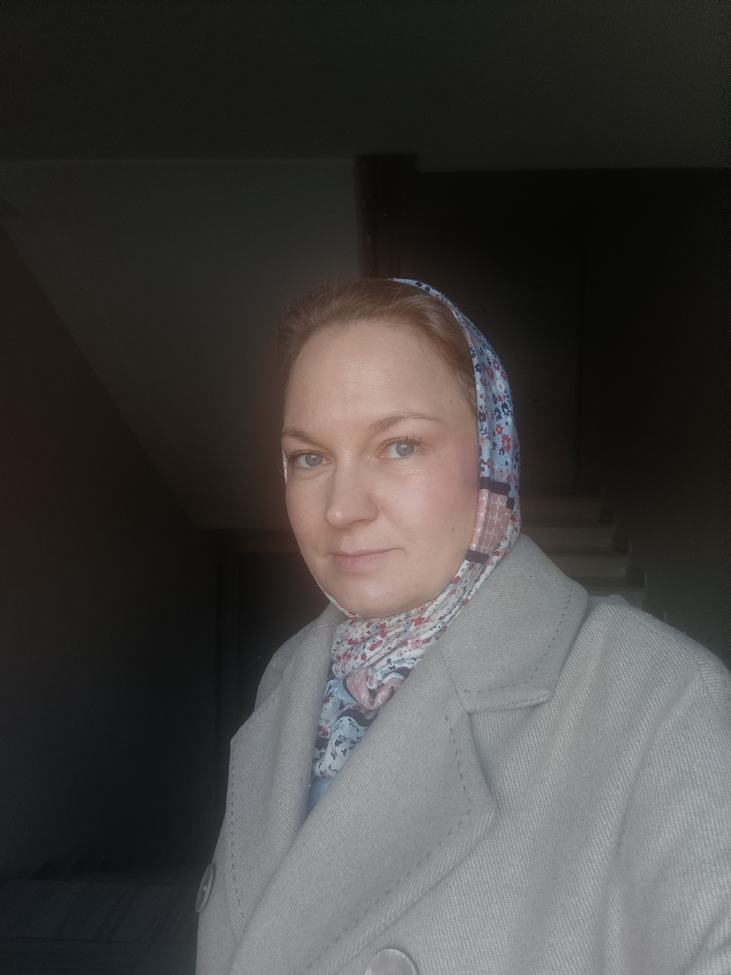 An adult female is depicted in a close-up, chest-up portrait within an indoor setting in the city of Марковский, Russia. She possesses fair skin, light-colored eyes, and light reddish-brown hair, partially visible at the forehead. Her expression is neutral, and she looks directly at the camera. She is wearing a light-colored, possibly beige or light grey, textured coat or jacket with a visible button on the left side. A multi-patterned headscarf, featuring blue, pink, red, and white hues in a floral or abstract design, covers her head and is secured around her neck. The background is dark and out of focus. On the right side of the subject, light-colored stairs are partially visible, indicating a stairwell or multi-level interior. The lighting appears to originate from the front-left, illuminating the subject's face and upper body against the darker environment. There are no other individuals or active interactions visible in the frame.