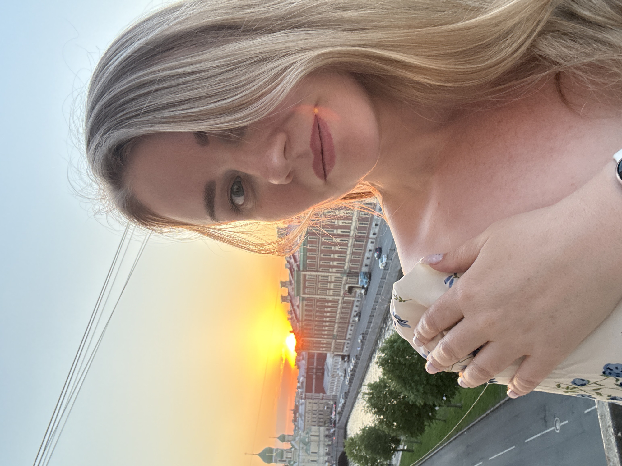 A vertically oriented close-up shot depicts a fair-skinned woman with blonde hair against a backdrop of a cityscape during sunset. The woman, positioned in the upper portion of the frame, appears to be holding the camera at an angle. Her expression is pleasant.
The background features a cityscape dominated by traditional Russian architecture, including red-brick buildings with ornate detailing. In the center of the frame, the sun is setting, casting a golden glow over the scene. A river or canal runs through the city, with trees and green spaces visible along its banks. A street with parked and moving vehicles can also be seen.
The geographical location is Saint Petersburg, Russia. The prominent red-brick building and sunset location are characteristic of Saint Petersburg. The angle of the photograph is such that the woman and the cityscape are captured in the same frame, creating a portrait with a scenic background.