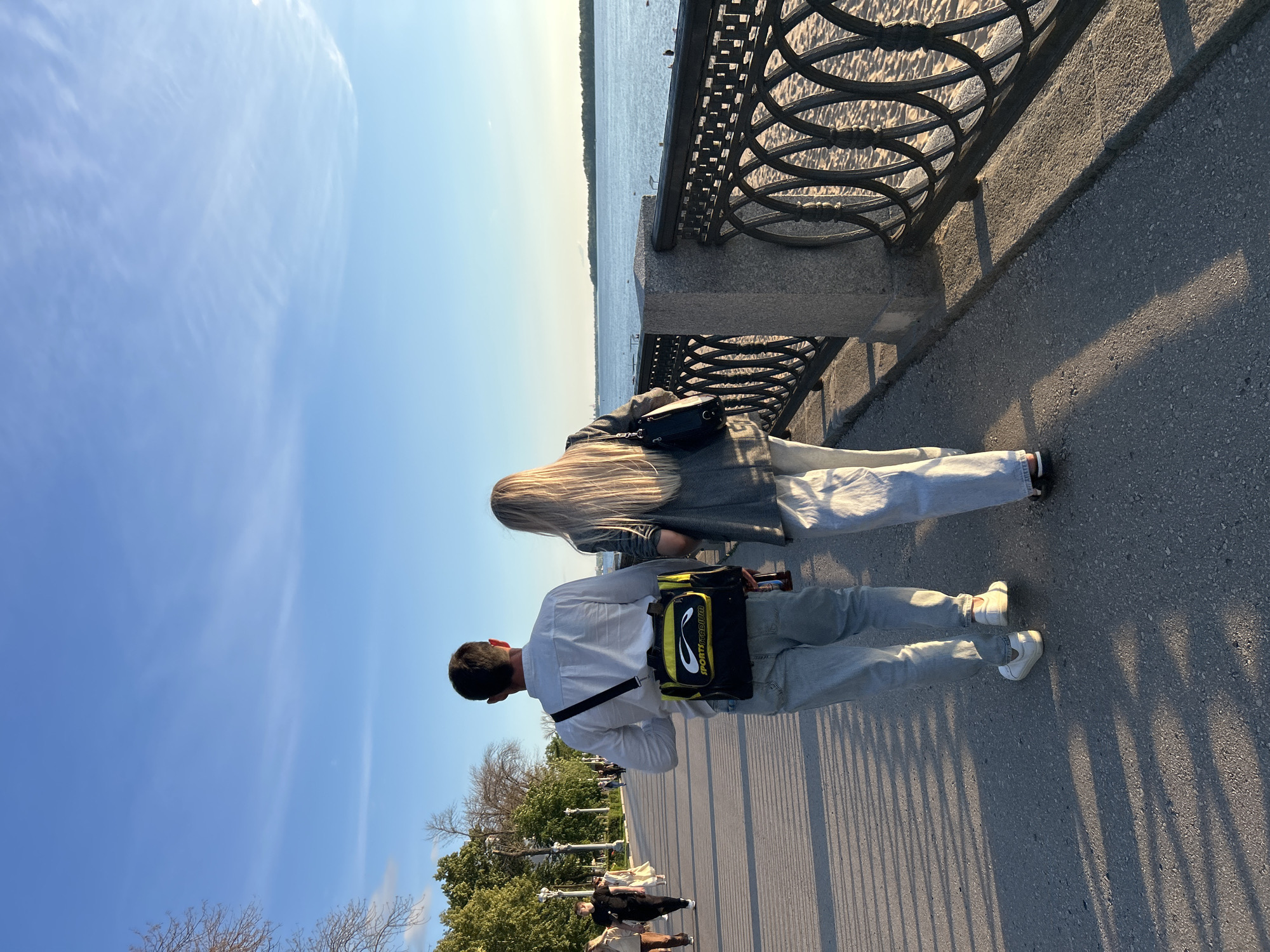 This is a vertical, outdoor photograph of two people standing on a paved walkway next to a body of water, possibly a river or canal. The scene is illuminated by bright sunlight, suggesting it is a clear day, likely late afternoon or early evening given the long shadows cast on the walkway.
In the foreground, a man is seen from behind, wearing a light-colored shirt, light-colored pants, and white sneakers. He has a black and yellow sports bag slung over his shoulder. To his left and slightly ahead, a woman with long blonde hair is also seen from behind. She is wearing a grey jacket over a light-colored top and light-colored pants, and a black purse is visible on her shoulder. They appear to be walking or standing along the waterfront.
Behind the people, a decorative metal railing runs along the edge of the walkway, separating it from the water. The water is calm and reflects the blue sky. In the distance, the opposite bank of the water is lined with trees. The sky is a vibrant blue with a few wispy clouds.
Further down the walkway, other people are visible in the distance, as well as trees and lampposts, suggesting a park or promenade area. The overall atmosphere seems relaxed and pleasant, typical of a leisurely stroll on a nice day. No specific emotions are overtly displayed by the subjects, but the scene evokes a sense of calm and enjoyment of the outdoors.
The word "SPORT" is visible on the yellow section of the bag carried by the man.
The image is oriented vertically.