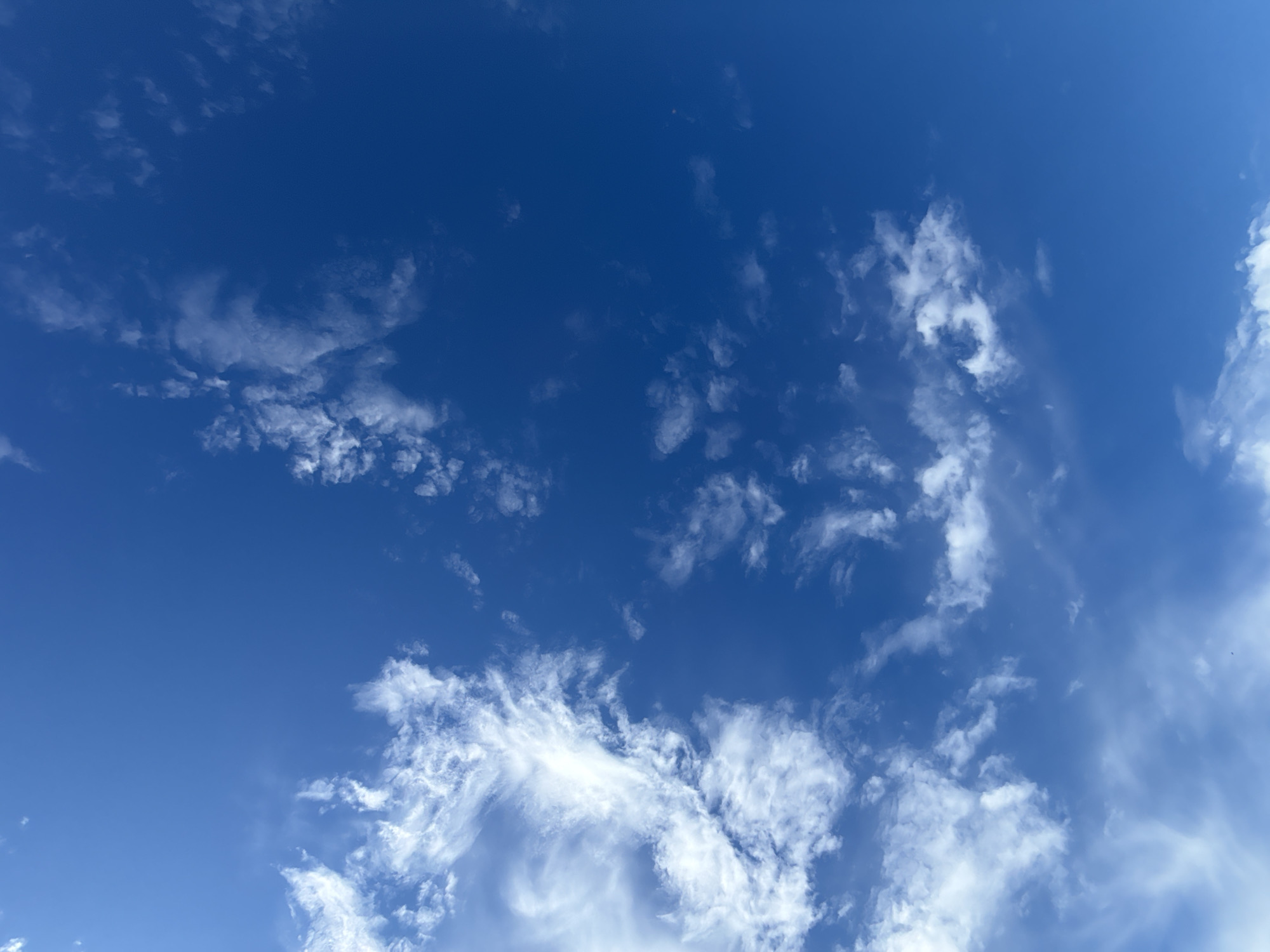 The image shows a clear blue sky with scattered white clouds. The clouds are wispy and appear to be high in the atmosphere. The lighting suggests it is daytime. No people, subjects, objects, or actions are visible in the scene, other than the natural elements of the sky and clouds. The location is identified as Hostomel, Ukraine.