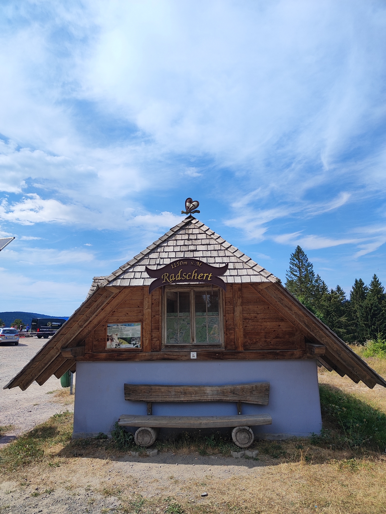 A rustic wooden hut with a grey base is situated outdoors on a bright, sunny day under a sky with scattered clouds. The hut has a shingled A-frame roof topped with a decorative metal weather vane shaped like a heart. A brown wooden sign on the roof reads "1150m ü. M. Radschert". Below the roofline, there is a window with a grid pattern. A wooden bench is placed in front of the grey base. To the left of the bench, a sign with images of wolves and text in German is mounted on the hut. In the background, there are trees and a distant view of hills. Several cars are parked to the left of the hut, and a black van with "SÜDHOLZ KAISER" printed on its side is visible. The ground is covered with dry grass and gravel.