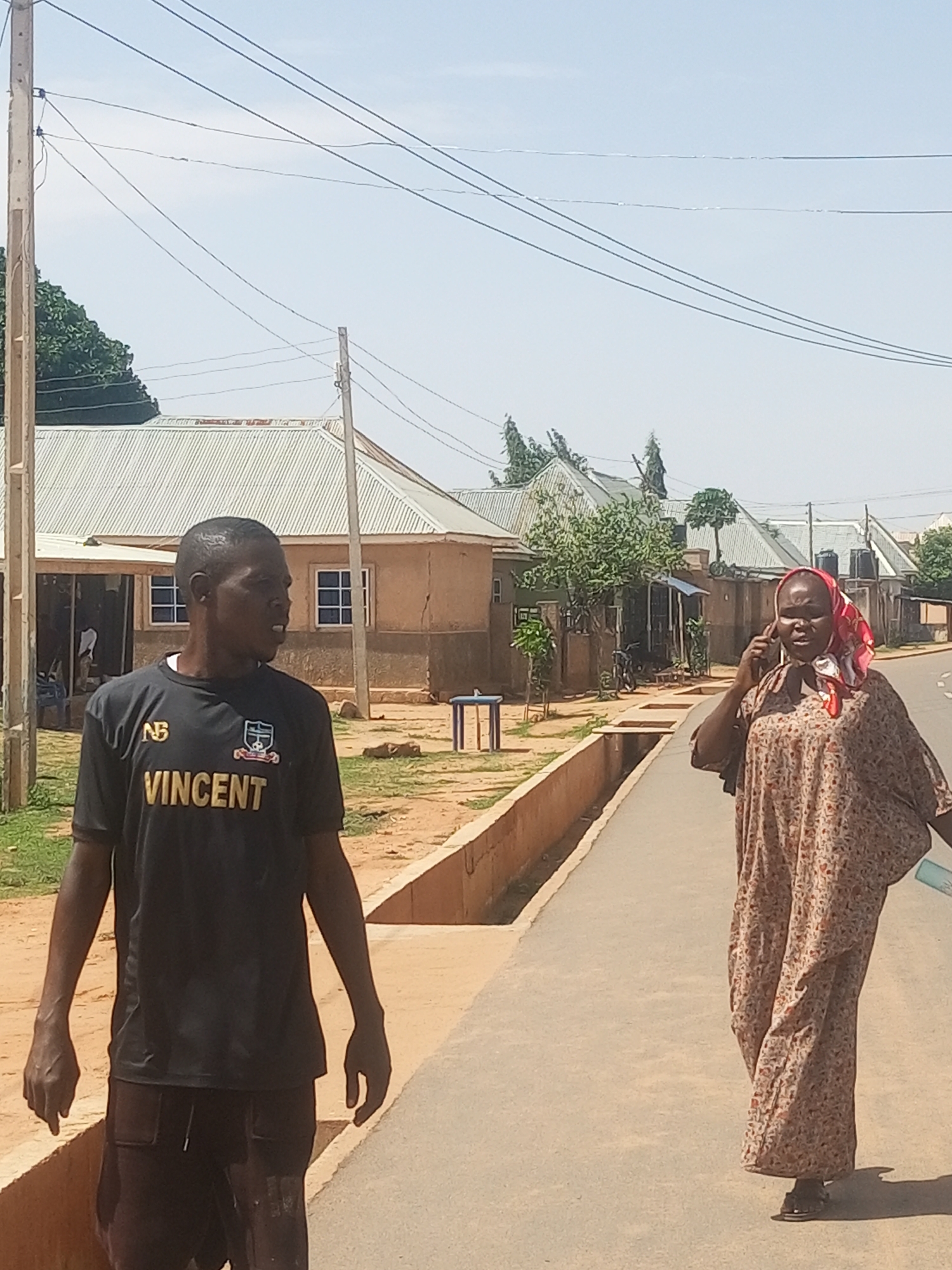 This daytime image, set in Tempure, Nigeria, features two individuals on a paved road with houses in the background. The sky is bright and clear, indicating sunny weather, likely late morning or early afternoon given the short shadows.
In the foreground, a man with short dark hair stands facing slightly right. He wears a black short-sleeved t-shirt adorned with a gold "NB" logo on the left chest and the name "VINCENT" printed in gold capital letters below it, alongside a small crest. He has dark-colored shorts or trousers.
Further down the road and to the right, a woman walks towards the viewer. She is dressed in a long, loose-fitting, light-colored dress with a subtle pattern, possibly floral. Her head is covered with a red and white patterned headscarf. She holds a dark object, likely a mobile phone, to her right ear, appearing to be in conversation. Her left hand holds a light blue object, possibly a bottle or a small bag.
The background shows a residential area with several single-story houses featuring light-colored corrugated metal roofs and earth-toned walls. Power poles and numerous overhead power lines are visible against the clear sky. Sparse trees and green foliage are scattered between the buildings. A concrete drainage ditch runs alongside the road on the left. The overall scene depicts a typical street in a semi-urban or rural Nigerian settlement.