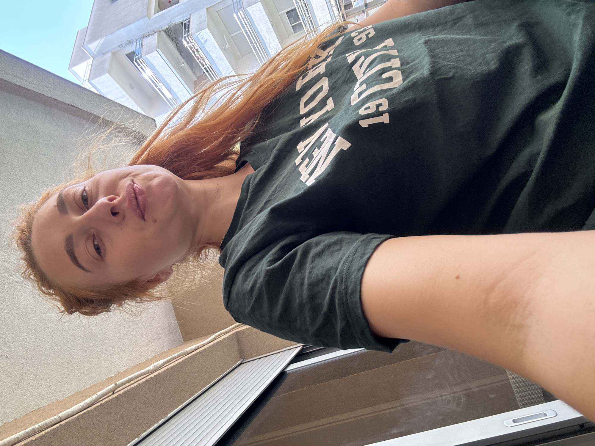 A woman with red hair is taking a selfie from a low angle, looking down into the camera. She is wearing a dark green t-shirt with white writing. The building behind her is likely located in Budva, Montenegro. A sliding glass door is visible below her.