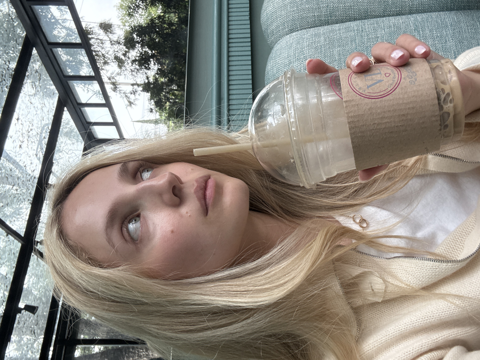 A young woman with blonde hair and fair skin is holding a plastic cup with a straw and coffee. She is wearing a cream-colored cardigan over a white shirt and a gold necklace. Her nails are painted pink. The background shows a teal-colored upholstered seat and a glass ceiling with a black frame. The scene suggests an outdoor cafe or seating area in Mexico City, Mexico.