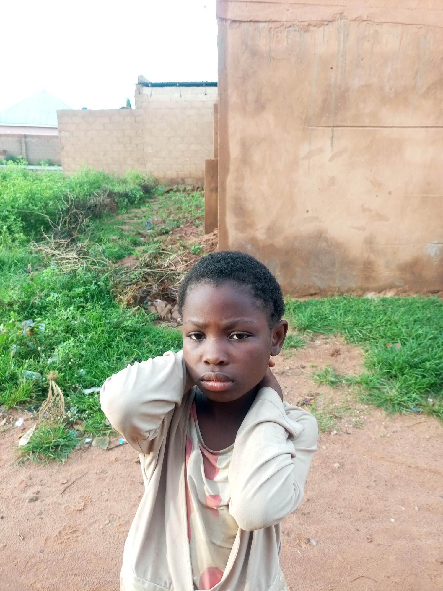A young Black child, who appears to be female, stands in the foreground of the image. She has dark, short hair, dark eyes, and medium-dark skin. She is wearing a light-colored, long-sleeved shirt over a patterned garment. Her hands are clasped behind her neck. The background consists of a dirt path, an area of dense green grass with scattered debris, and a light brown wall of a building. In the distance, there are more brick structures and a light-colored roof with a blue or green hue. The scene appears to be outdoors in a dusty, possibly undeveloped urban area in Tempure, Nigeria. The lighting suggests it is daytime, and the sky is overcast.