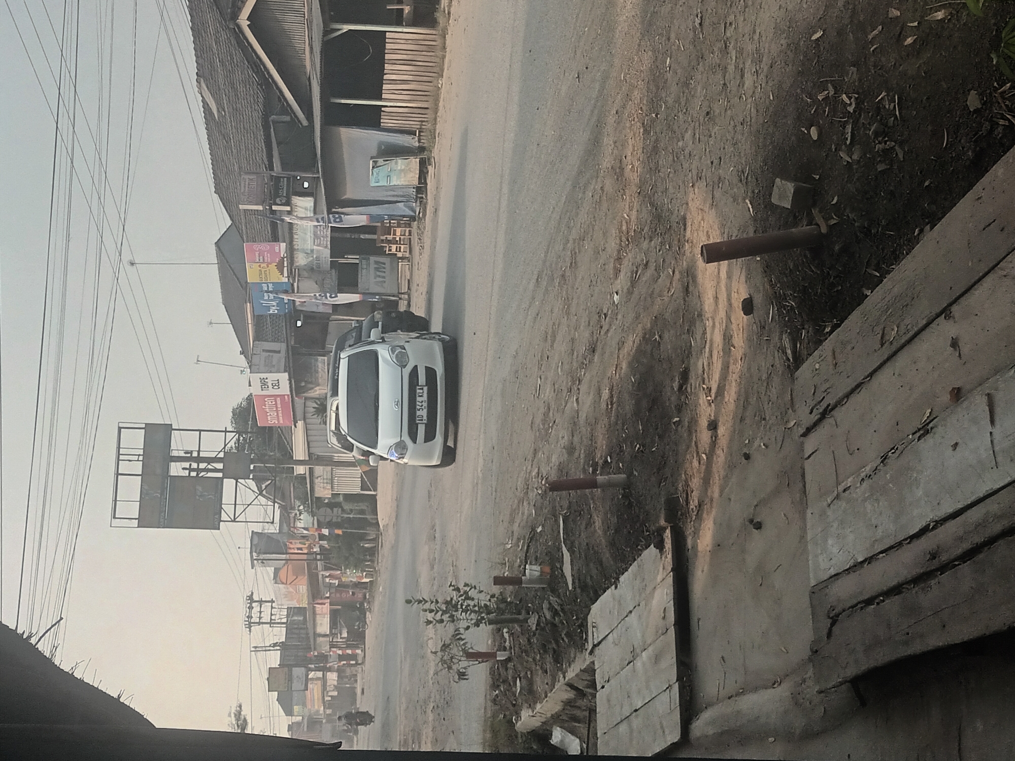 The image depicts a street scene in Mulyo Asri, Indonesia, with a focus on the left side of the road. A white sedan, license plate AD 544 KLA, is driving down the paved road. The road is flanked by uneven dirt verges. On the left verge, several wooden planks are laid out, suggesting a temporary walkway or construction material. There are also small, newly planted trees with red and white markers. Further up the road, buildings with various signs, including "Smartfren" and "Tempe Cell", are visible. A tall advertising billboard is positioned prominently. In the distance, several motorcycles and a person on a motorcycle can be seen. The sky is overcast and hazy. The right side of the road features a row of buildings with corrugated iron roofs and some covered stalls. An ATM machine is visible next to one of the stalls. The overall environment appears to be a semi-urban or rural commercial area.