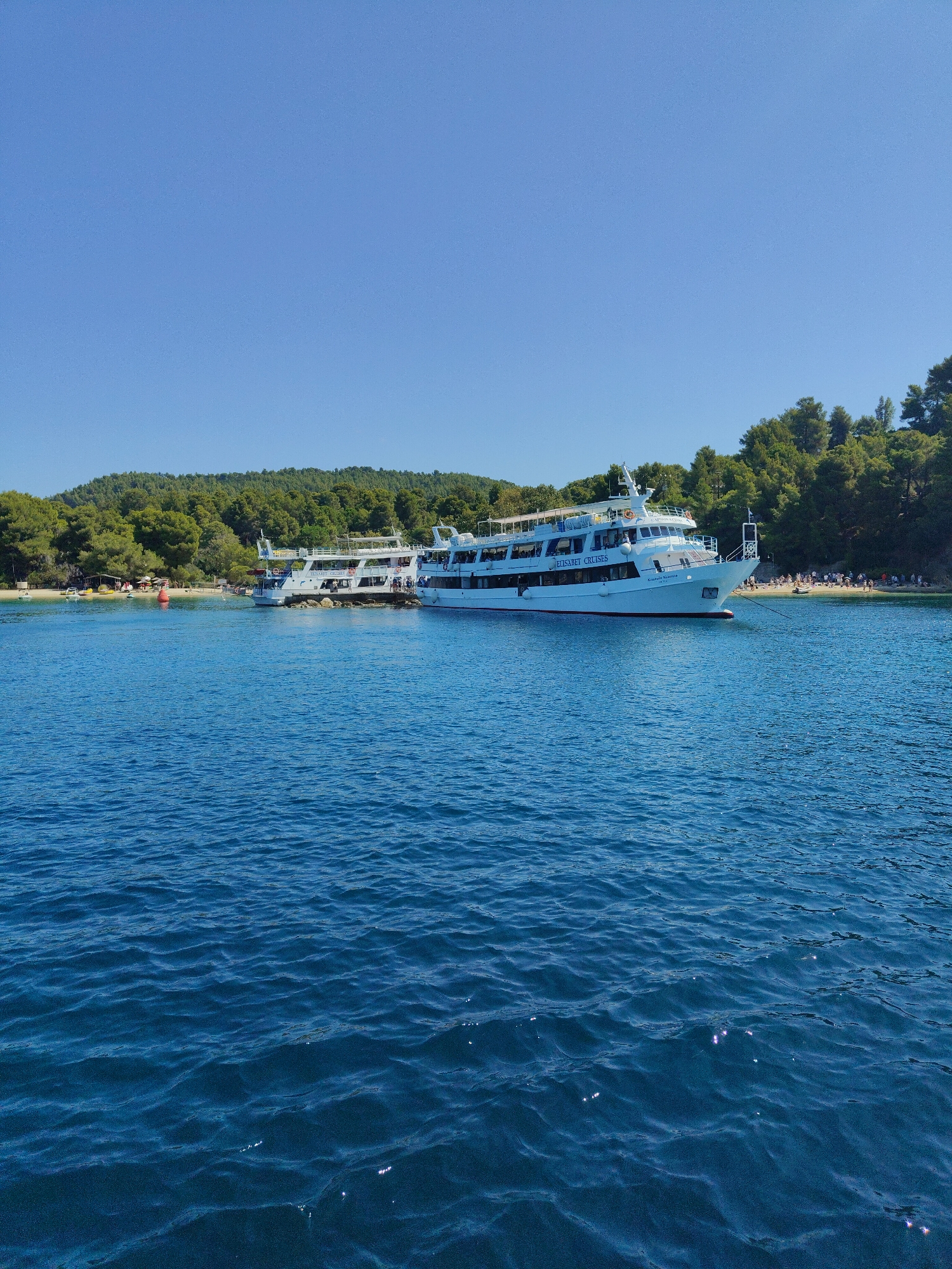 This is a wide-angle, eye-level shot taken from the water, looking towards a shoreline with trees and boats. In the foreground, the deep blue sea is choppy with visible ripples and sparkles from the sunlight. Two white passenger boats are moored in the bay, one significantly larger than the other. The larger boat, which appears to be a cruise or ferry vessel, has multiple decks and a white hull. The text "ELISABET CRUISES" is visible on its side. The smaller boat is docked behind it. Along the shoreline, a sandy beach is visible with scattered people. Behind the beach, a dense green forest covers a hillside. The sky above is a clear, bright blue. The scene suggests a sunny day at a coastal location in Greece.