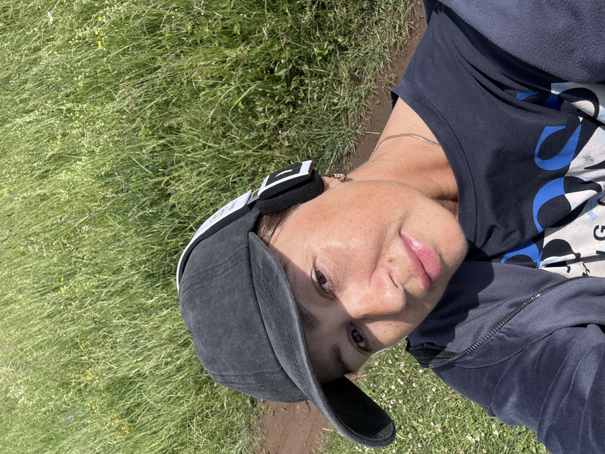 A medium shot selfie shows a woman wearing a dark baseball cap and headphones. She has on a dark blue t-shirt and a dark blue zip-up jacket. The woman is looking down and to her right with a slight smile. Her hair is dark brown and pulled back. The background consists of tall green grass and some brown dirt. The location is the city of Kalinin, Russia.