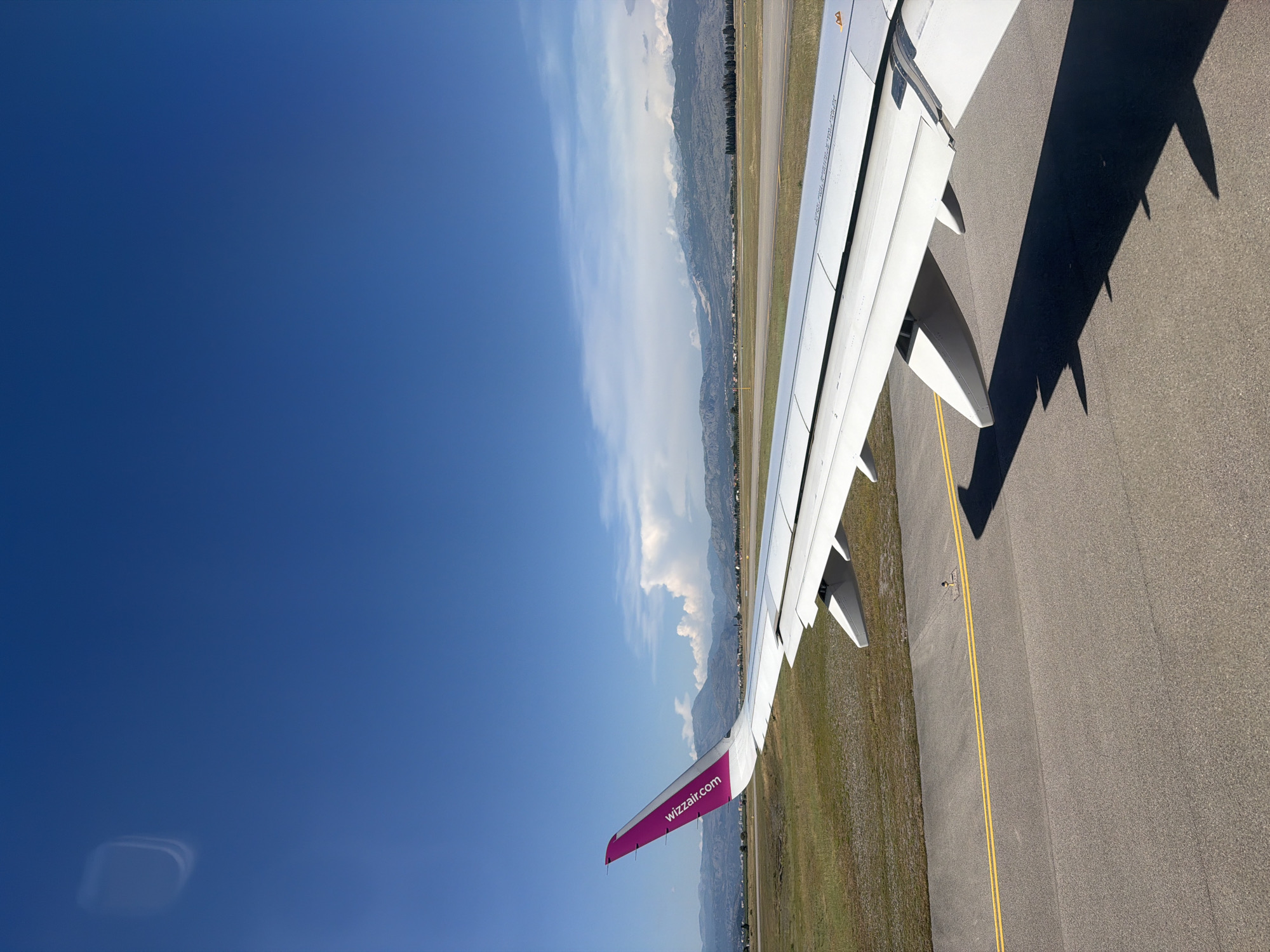 The image shows an airplane wing with the Wizz Air logo on the purple wingtip. The wing is positioned over an airport tarmac with yellow lines marking a runway. In the background, there are green fields, trees, buildings, and mountains under a clear blue sky with scattered clouds. The scene appears to be at the Podgorica Airport in Donja Cijevna, Montenegro. There are no people visible in the image. The airplane is either on the ground or taxiing, as indicated by the shadow of the wing on the tarmac.