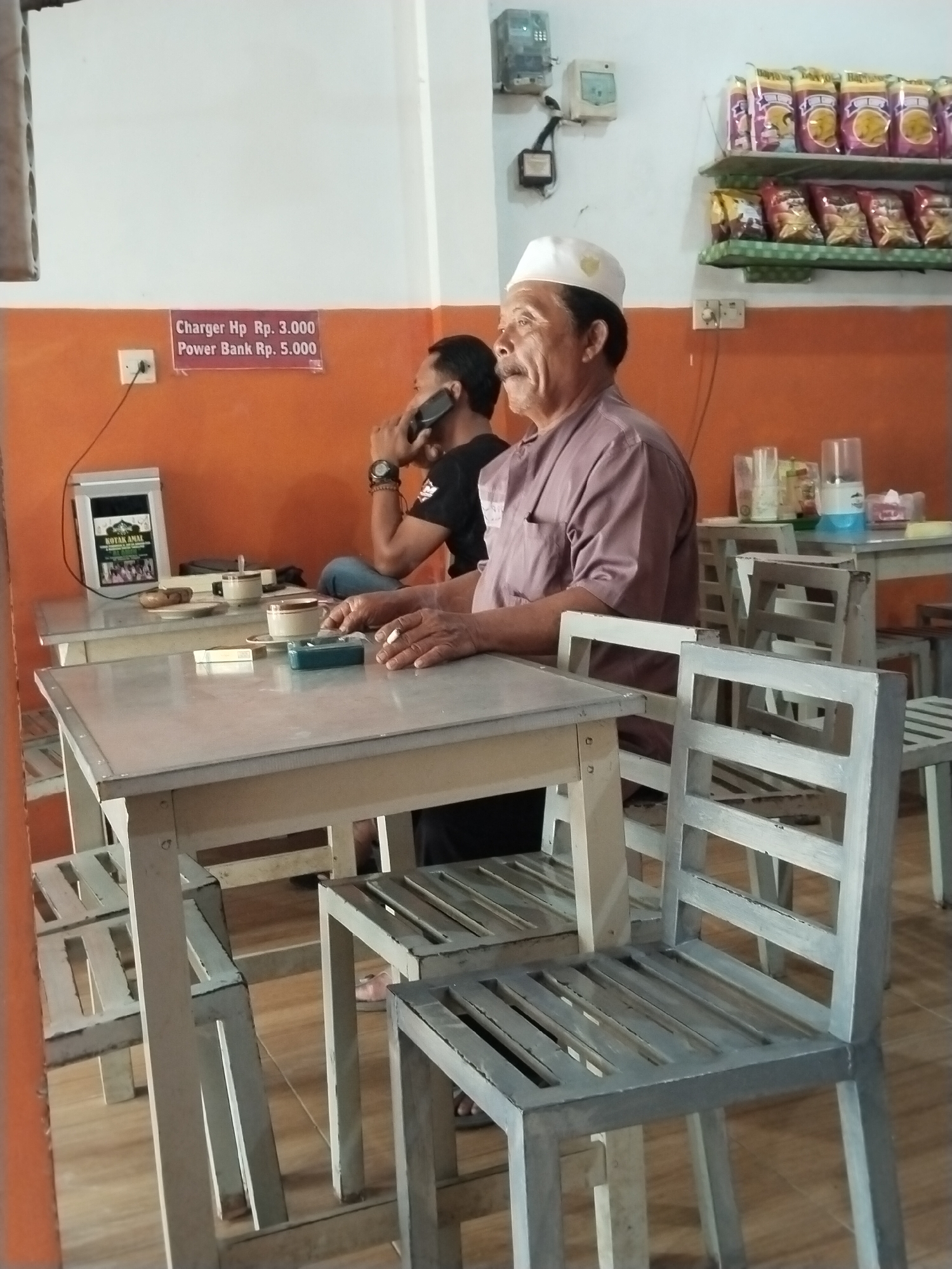 The image depicts an indoor scene, likely a small shop or eatery, with an orange-painted wall and white upper walls. In the foreground, a man wearing a white cap and a muted reddish-brown shirt is seated at a light-colored metal table, holding a cigarette. Another man, in the background, is seated at a similar table, talking on a mobile phone, wearing a dark shirt and blue jeans.
On the tables, there are cups and an ashtray. A sign on the wall reads "Charger Hp Rp. 3.000, Power Bank Rp. 5.000". Shelves on the upper wall display various bagged snacks. Electrical outlets and meters are visible on the wall. The furniture, including tables and chairs, appears to be made of light-colored metal. The floor is a wooden plank. The setting suggests a casual establishment in a city like Prigi, Indonesia, where such services and goods are commonly offered.
