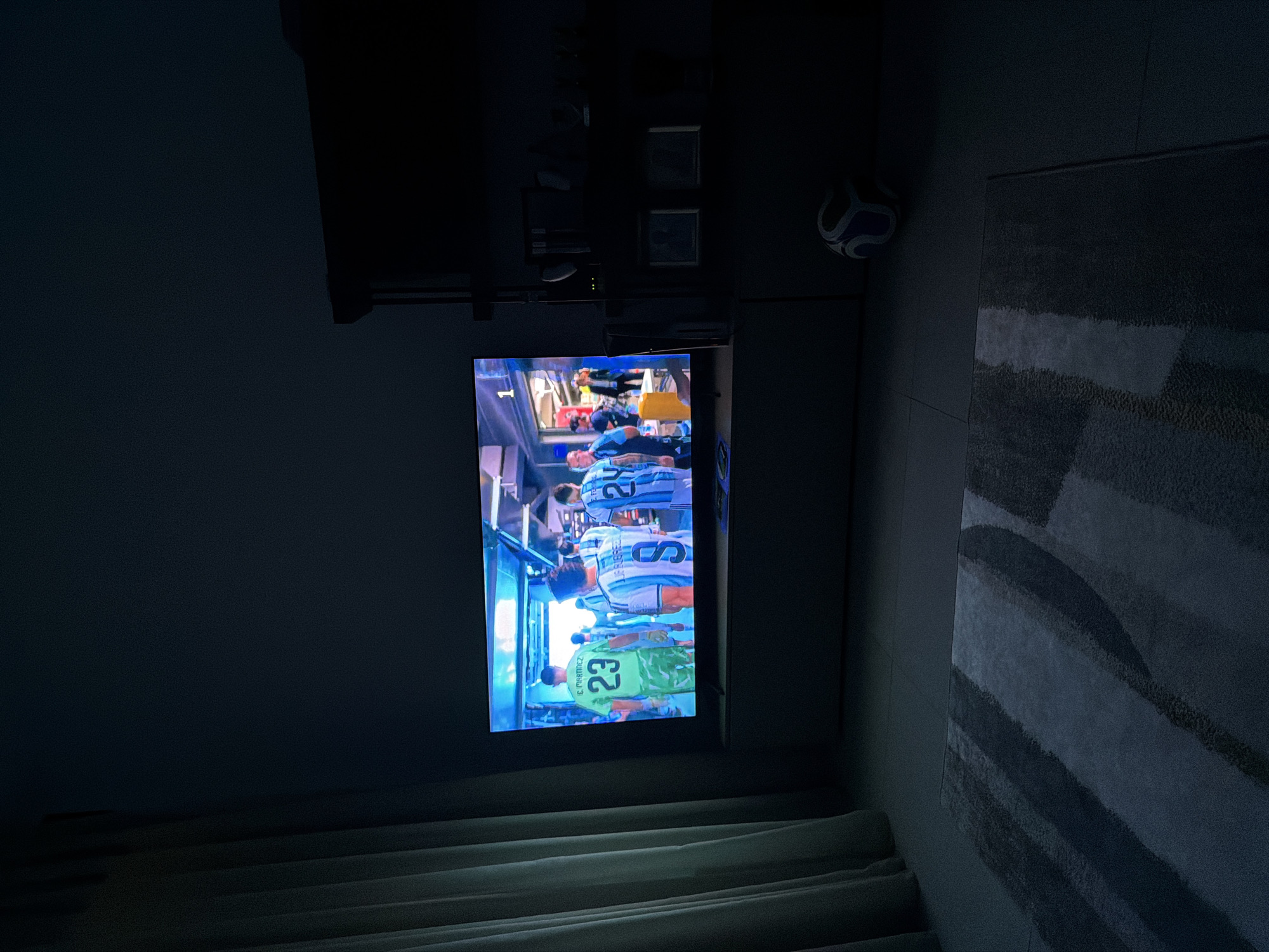 A television screen displays a soccer match, with players in uniform walking through a tunnel. The room is dimly lit by the television's glow, with a curtain on the left and a bookshelf on the right. A rug lies on the floor in front of the television, and a soccer ball is visible on the floor to the right. The setting appears to be a living room in Finestrat, Spain.
