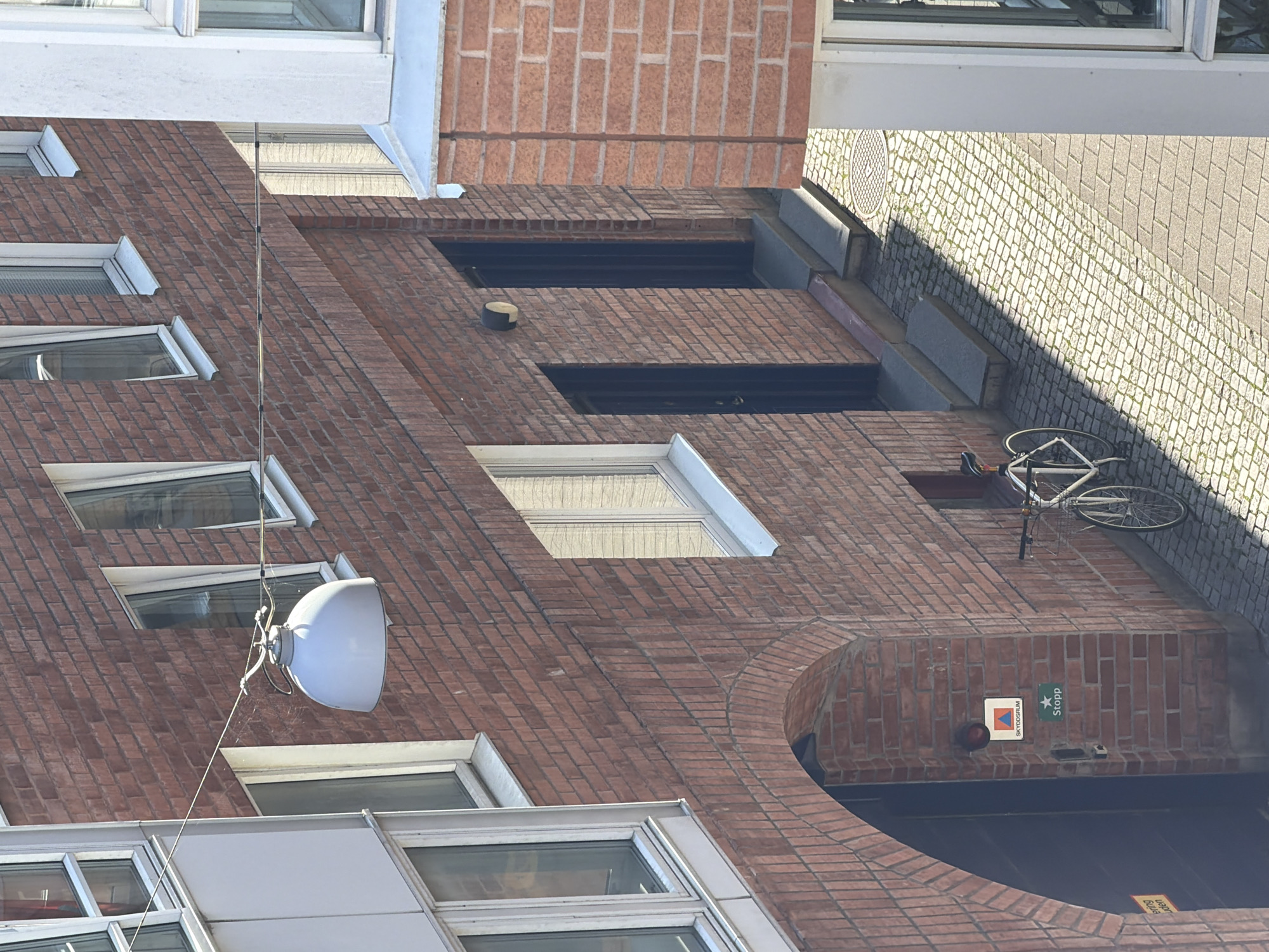 The image shows an exterior view of a building in Malmö, Sweden, constructed with red brick. There is a light fixture hanging from a wire strung across the building. A bicycle is parked near an arched entryway. Two dark doors are visible on the building, one with steps leading up to it. There are several windows on the brick facade, some with blinds. The ground is paved with cobblestones. The building appears to be a multi-story structure.