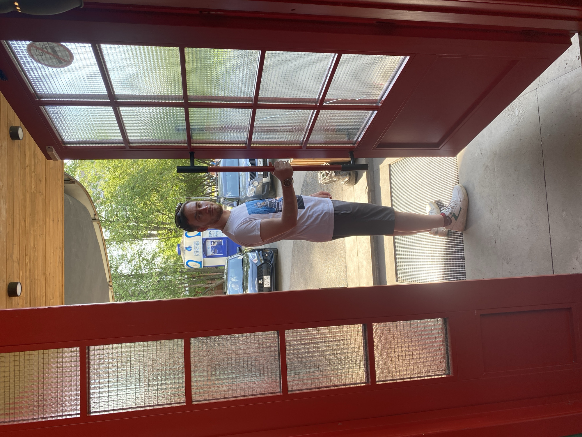 A man is photographed standing in an open, red, glass-paneled doorframe. He's wearing a white t-shirt, grey shorts, and white sneakers with red and green accents. He is holding onto the horizontal metal bar of the doorframe with his left hand and wearing a watch on his left wrist. The doorframe appears to be the entrance to a building. Outside, a blue car is visible along with signage in Russian, indicating the city of Izhevsk, Russia. The overall scene is outdoors with trees and daylight conditions.
