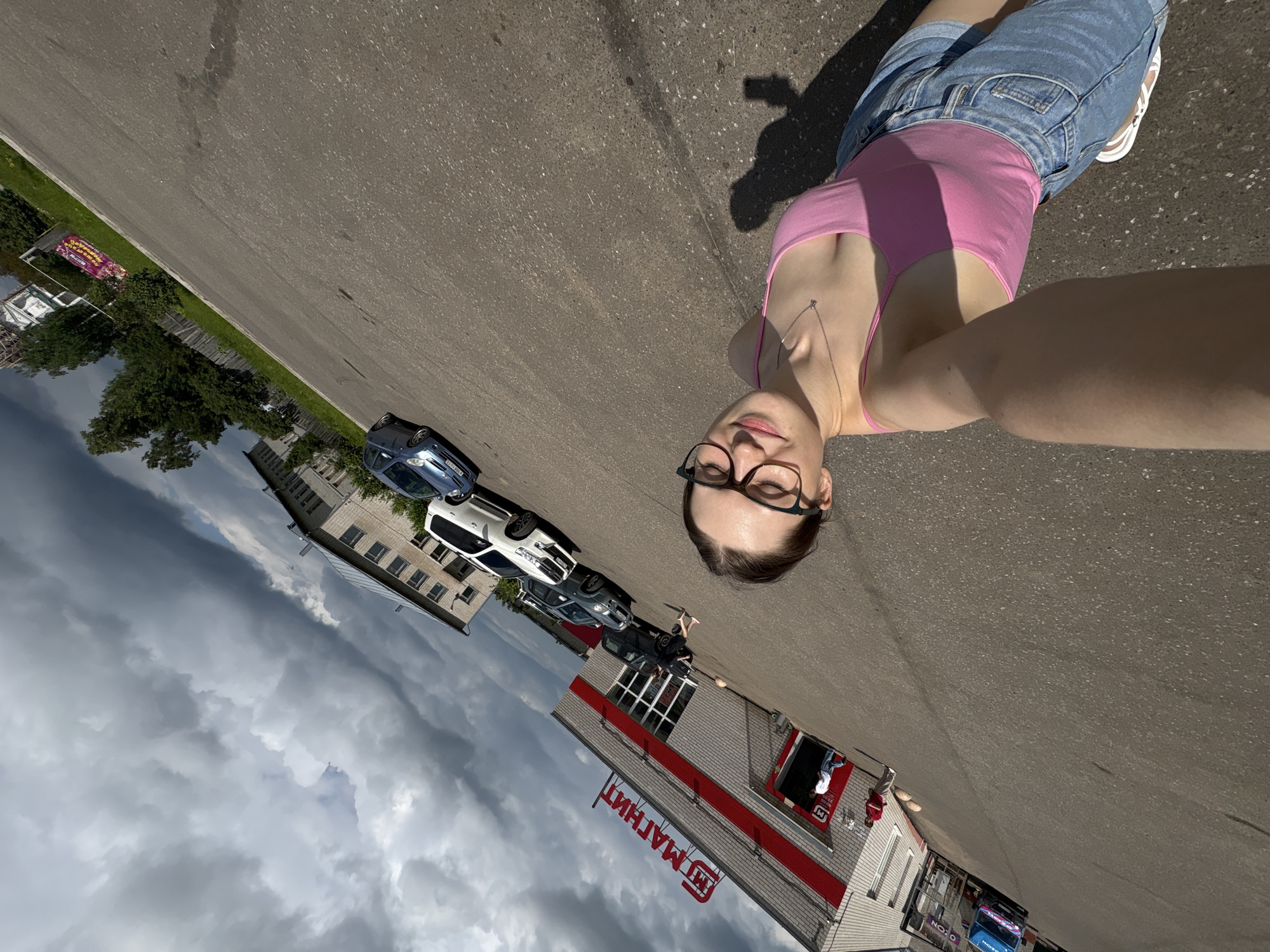 A low-angle, full-body selfie is taken outdoors in front of a "Magnit" grocery store in the city of Kstovo, Russia. The sky is heavily clouded, with dark storm clouds dominating the upper portion of the frame, contrasting with patches of blue sky. The store, a single-story building with a red and white facade and a prominent red "Magnit" logo, is visible on the left. Several cars are parked in front of the store, including a white SUV, a dark sedan, and a blue hatchback.
The subject, a young woman with glasses, is in the foreground, wearing a pink tank top and denim shorts. She is smiling and holding her phone to take the picture. Her left arm extends towards the camera, and a shadow of her and her phone is cast on the asphalt ground. The ground is paved asphalt, and a slight tilt is applied to the image, giving a dynamic perspective. In the background, a fence and trees are visible, with a distant building. The overall lighting suggests it's daytime, but the dark clouds indicate impending or recent rain.