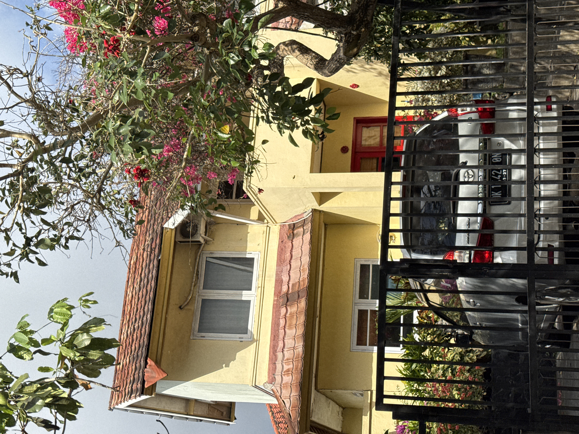 The image depicts the exterior of a two-story yellow house in Kuta Selatan, Indonesia. The house has a red-tiled roof and white-framed windows. A large tree with pink and red flowers is in the foreground, partially obscuring the house and a black metal fence. Behind the fence, a silver car is parked. The environment suggests a sunny day with some clouds.